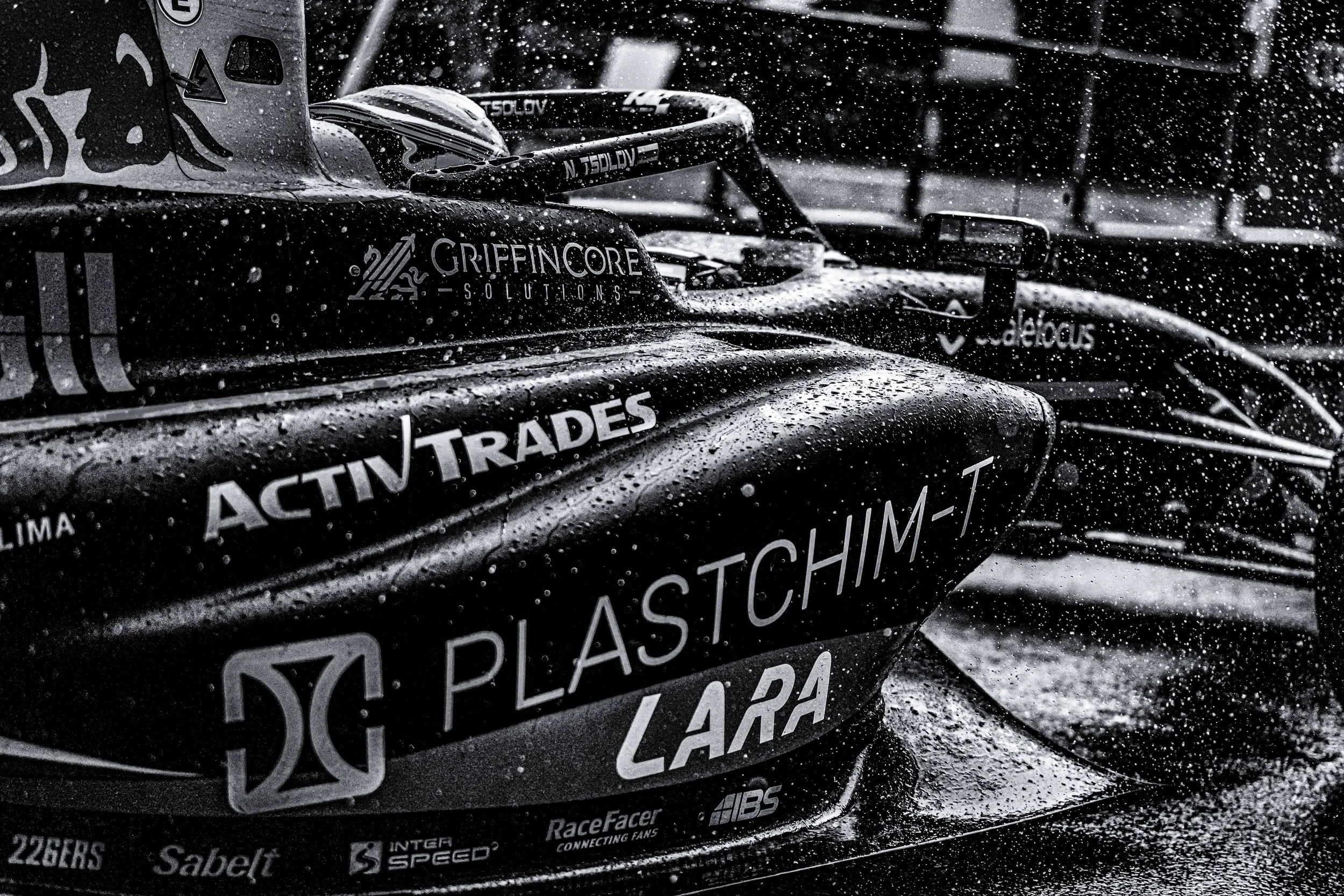 Close-up of a race car covered in rain droplets, showing various sponsor logos and decals on the body of the vehicle.