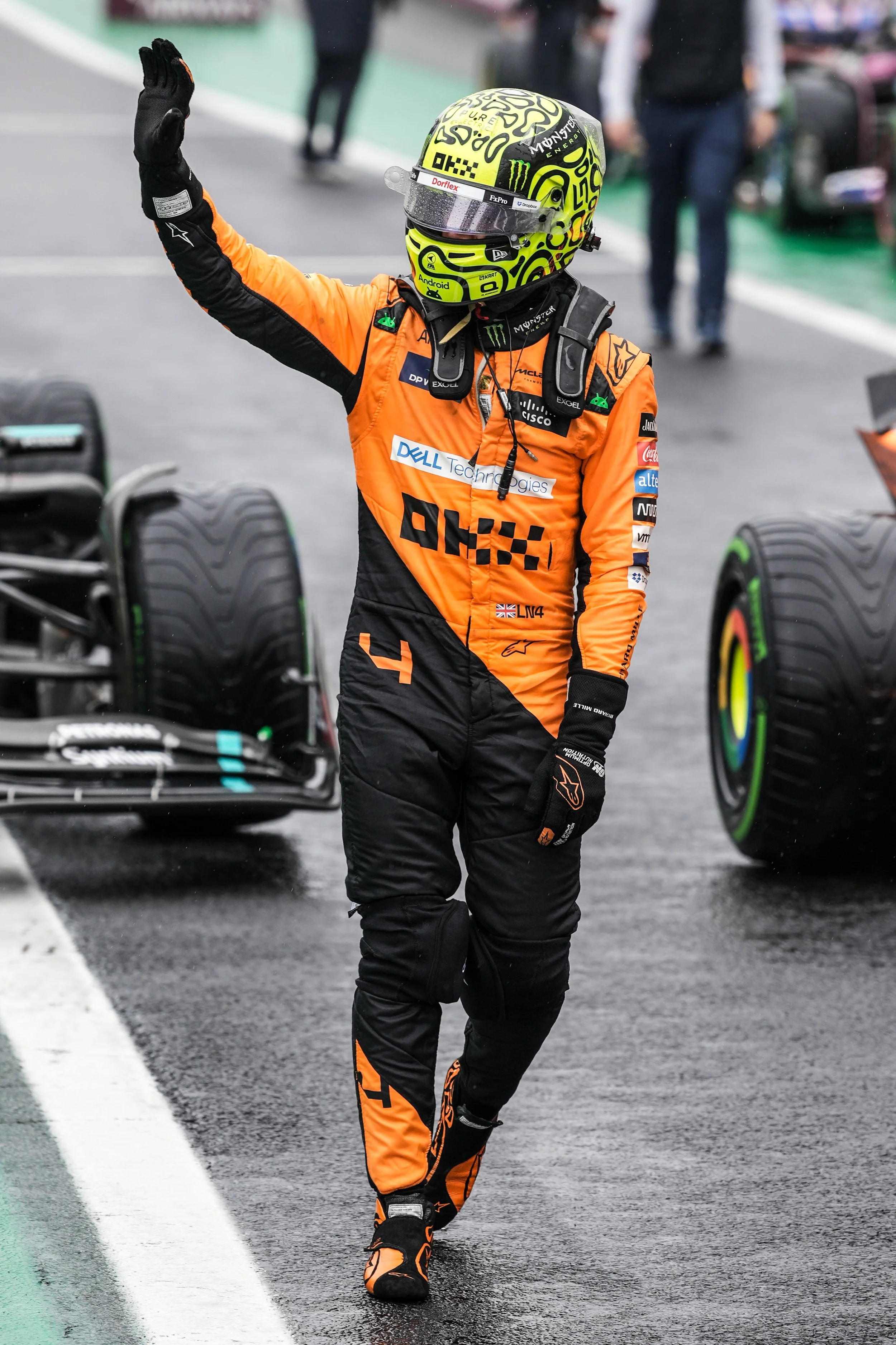 Lando Norris, waving to the crowd after a race, with racing cars in the background.