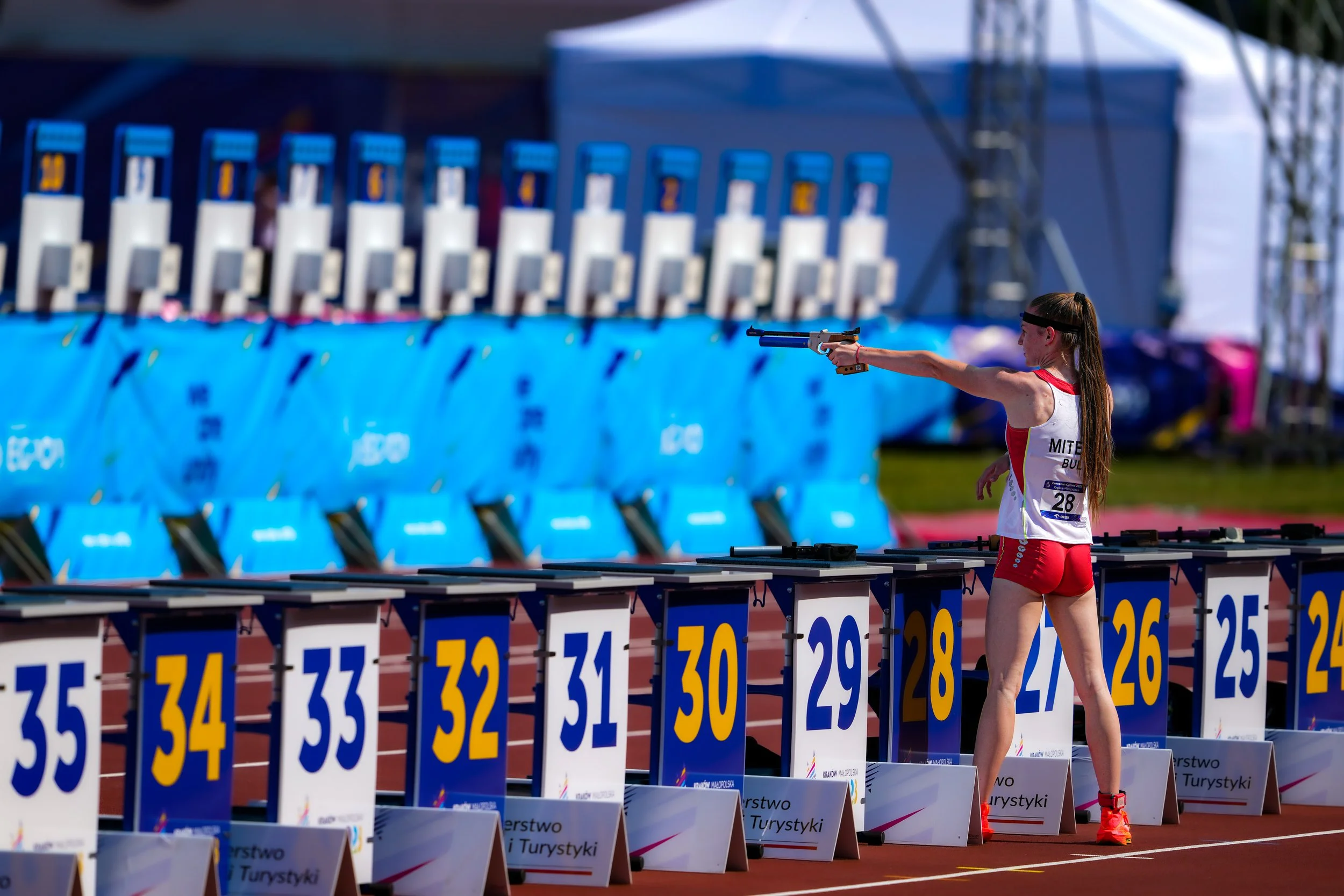 A female athlete in red shorts and a white tank top aiming a pistol at a shooting range during a competition.