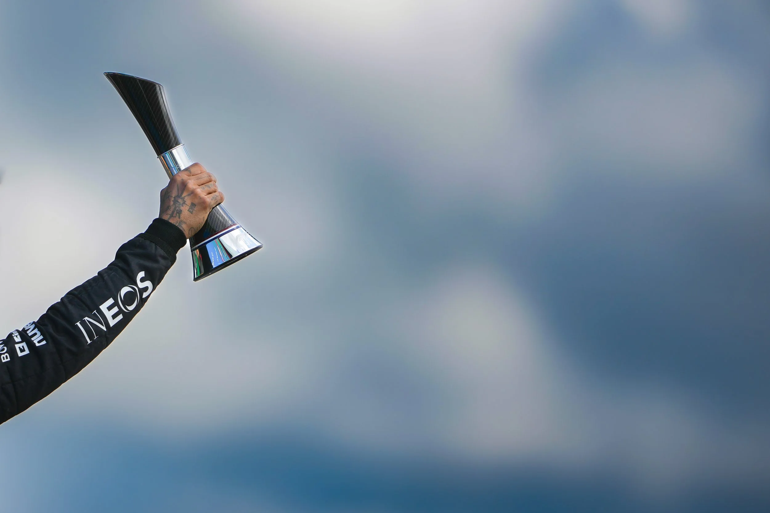 A person in a black racing suit holding up a trophy against cloudy sky.