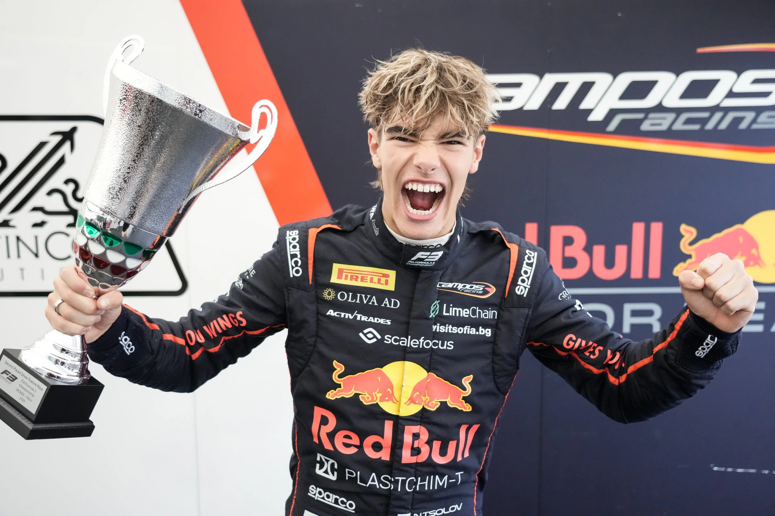 A race car driver celebrating on the podium with a trophy, wearing a racing suit with sponsor logos, including Red Bull and Pirelli, and making a victorious gesture with one hand.
