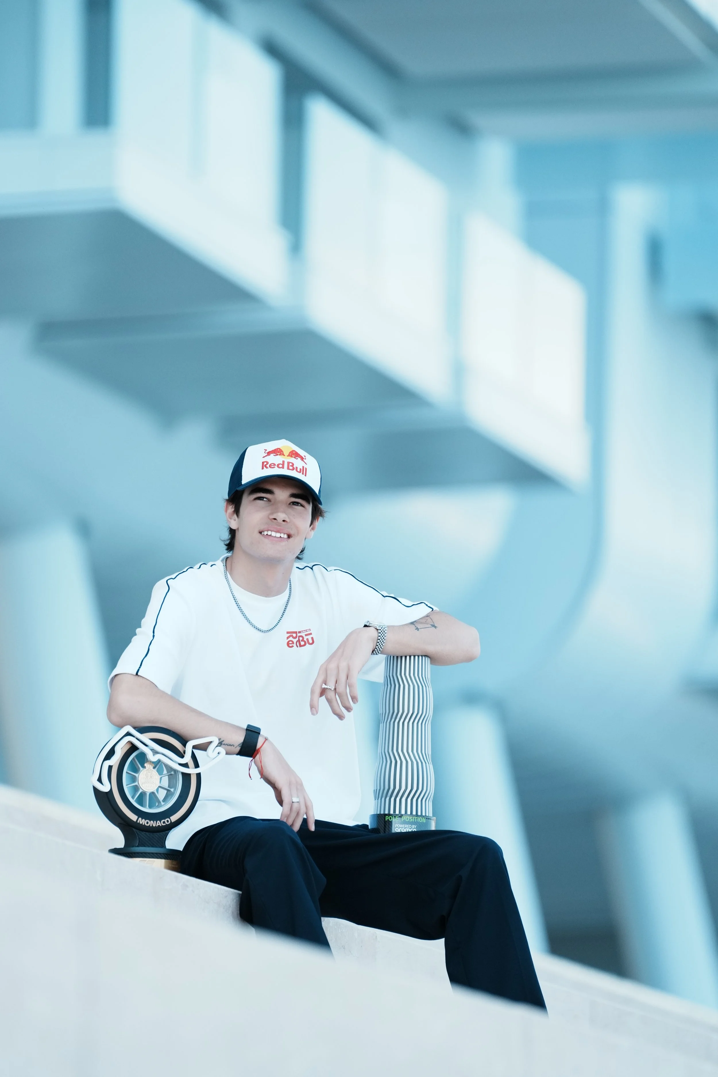 A young man wearing a Red Bull racing hat and white t-shirt with Red Bull logos, sitting on a ledge with a blue and white striped water bottle and a trophy, in a modern building with large glass windows.