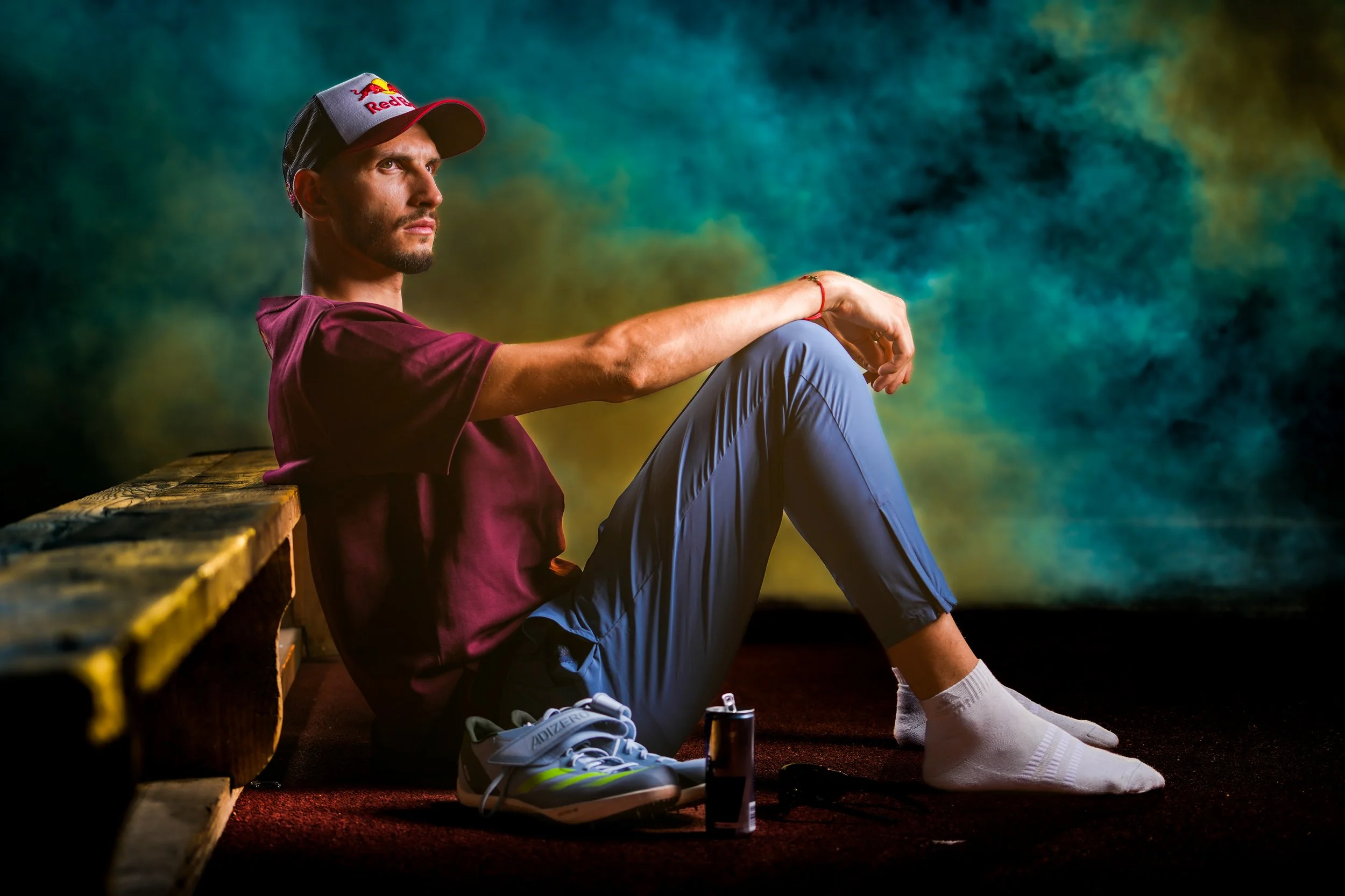 A man wearing a Red Bull cap, maroon sports shirt, gray pants, and white socks, sitting against a wooden bench with sneakers, a water bottle, and technology beside him, with a colorful abstract background.