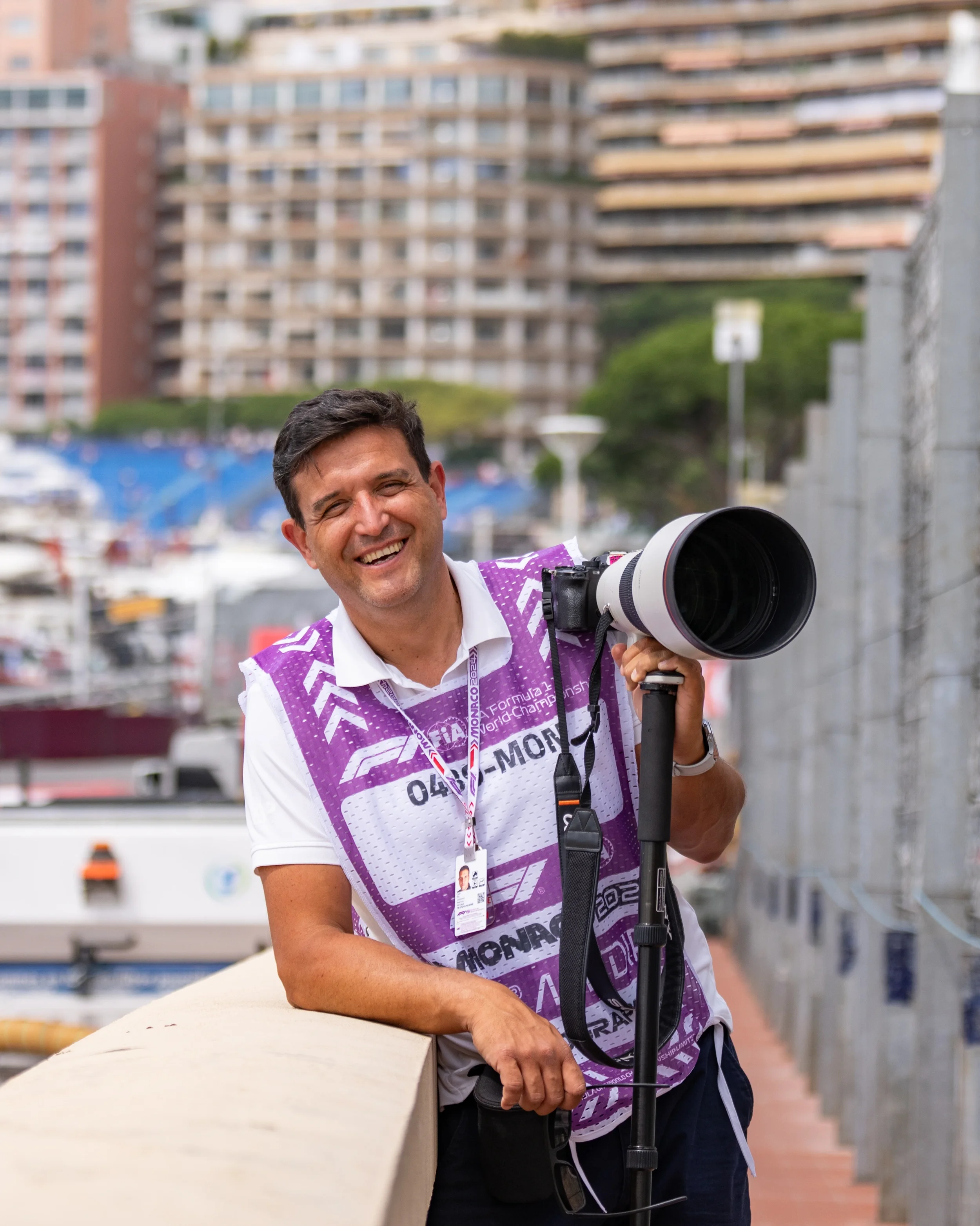 Lubomir Asenov smiling and holding a large camera lens on a tripod, standing outdoors near a marina with boats and high-rise buildings in the background.
