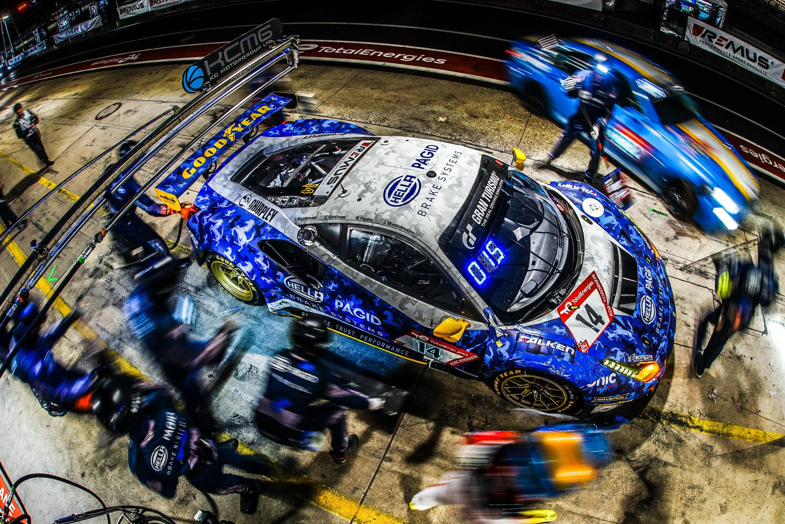 Race car in pit stop area with crew members working on it, surrounded by other race vehicles and personnel.