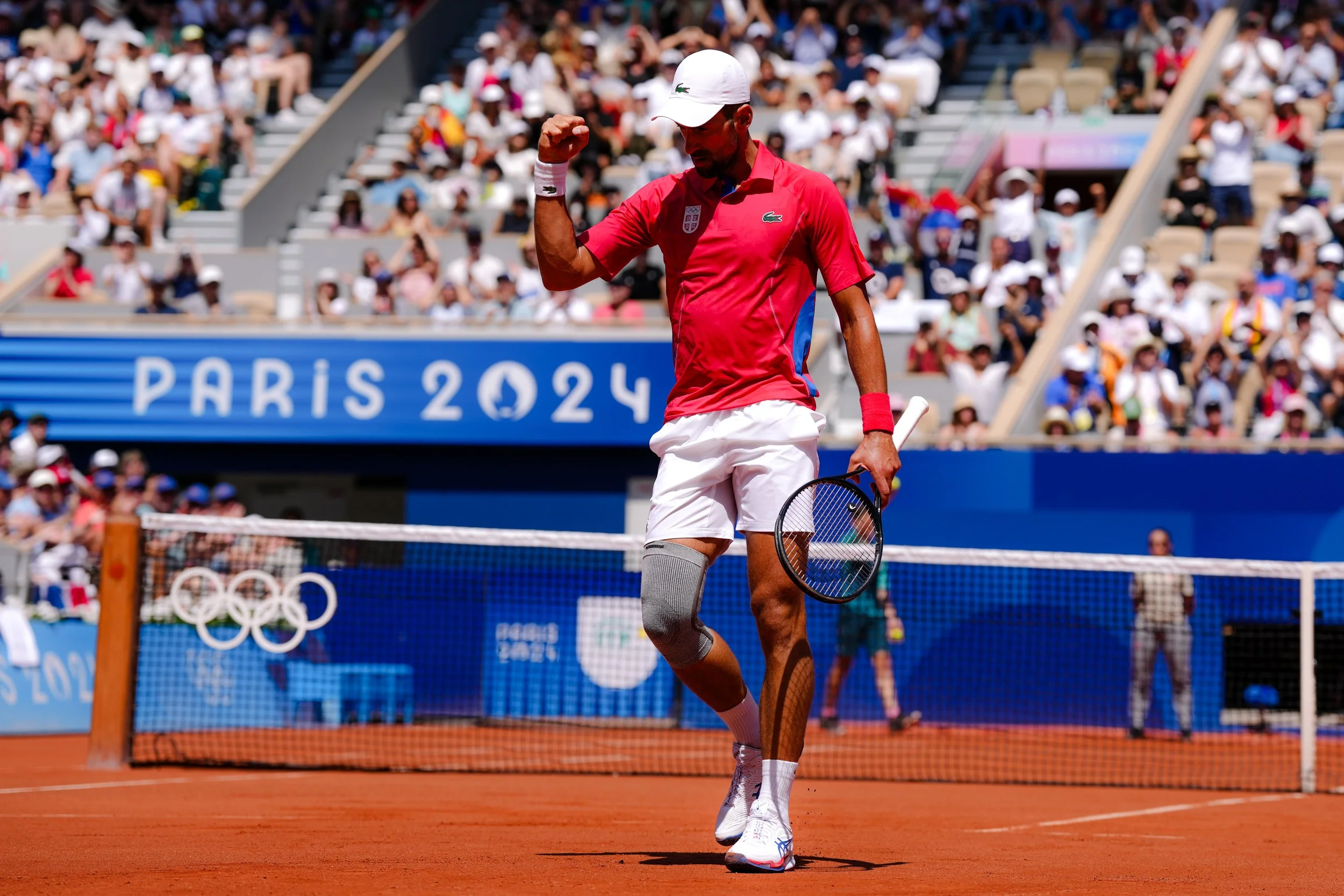 A male tennis player in a red shirt and white shorts flexing his right arm after winning a point during a match at the Tokyo 2020 Olympics, with spectators in the background and a banner that reads "Paris 2024".