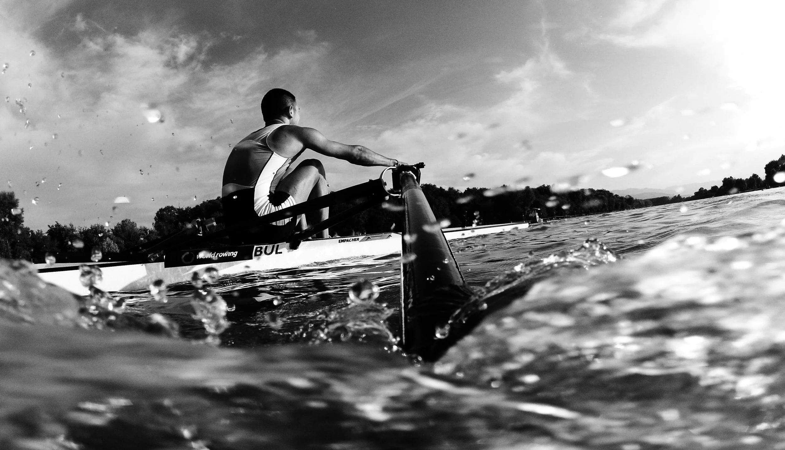 A person rowing a boat on a body of water, viewed from a low angle near the water surface, with water droplets and a scenic shoreline in the background in black and white.