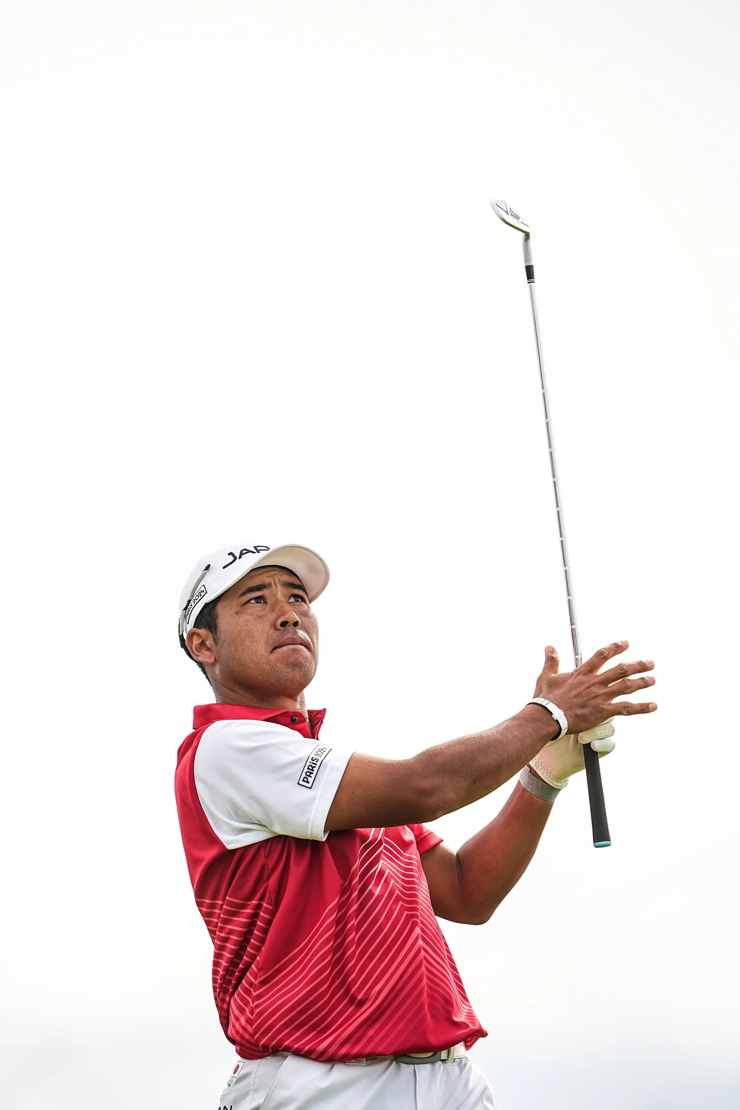 A male golfer in a red and white polo shirt, wearing a white cap, holding a golf club and focusing on the sky.