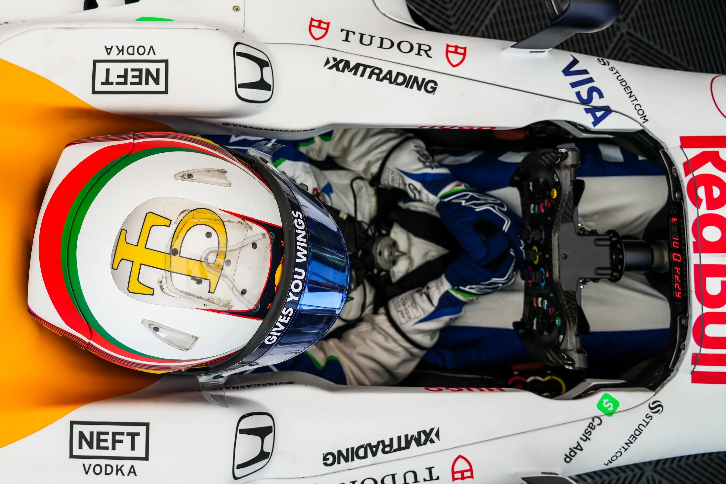 A race car driver sitting in a Formula 1 car, wearing a white helmet with red, green, and yellow accents, and a racing suit with blue and white colors. The car's cockpit is equipped with various controls and a digital display, and the car's body feat