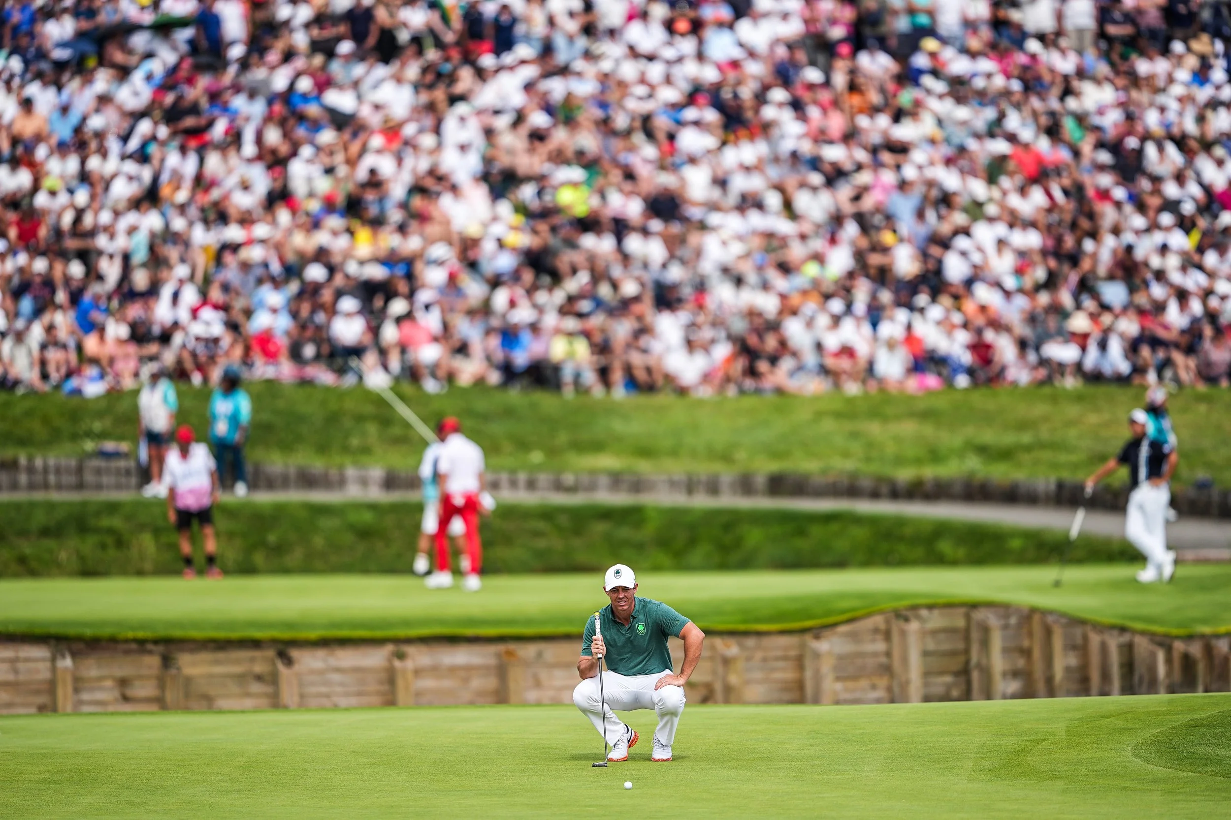 A male golfer crouching on the green with a golf ball in front of him, preparing for a putt, during a golf tournament with a large crowd of spectators in the background.