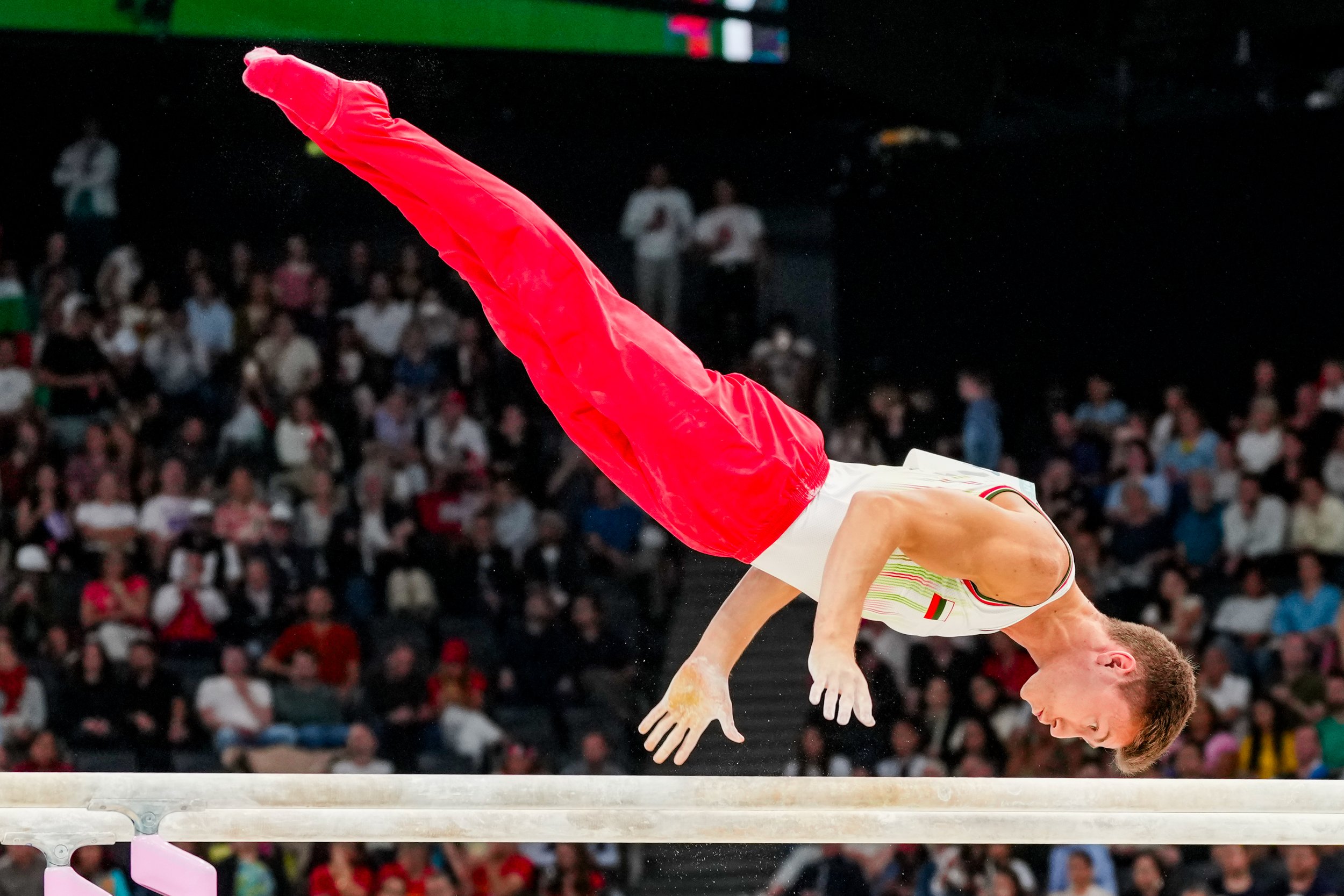 Male gymnast performing a vault on the horizontal bar during a competition, with an audience in the background.