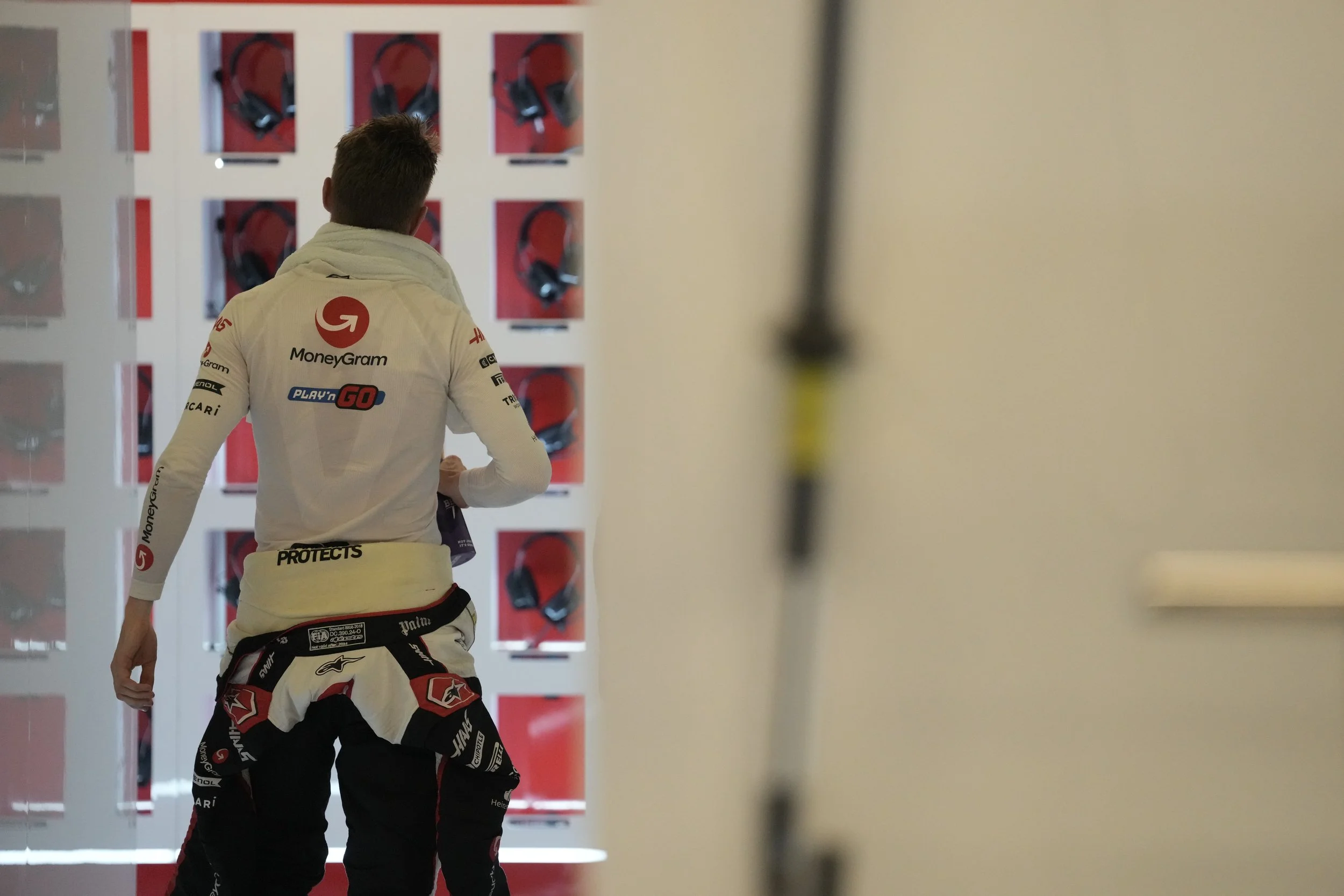 A race car driver in a white racing suit with sponsor logos, standing in front of a display case with racing helmets. The driver is seen from behind, with short hair, and appears to be preparing or inspecting something.