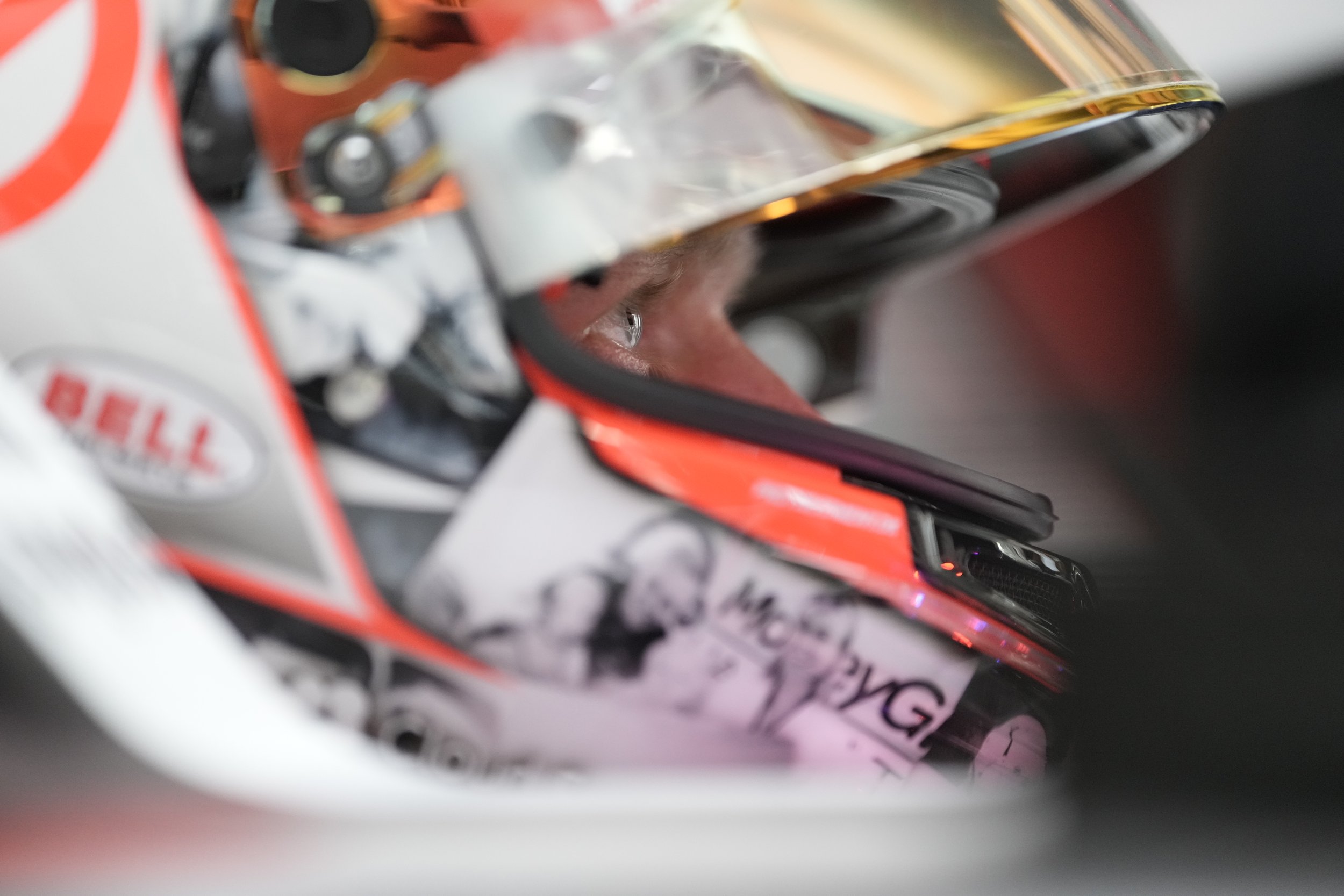 Close-up of a race car driver wearing a helmet with the visor lifted, revealing the eyes, inside a racing helmet with various sponsor logos.