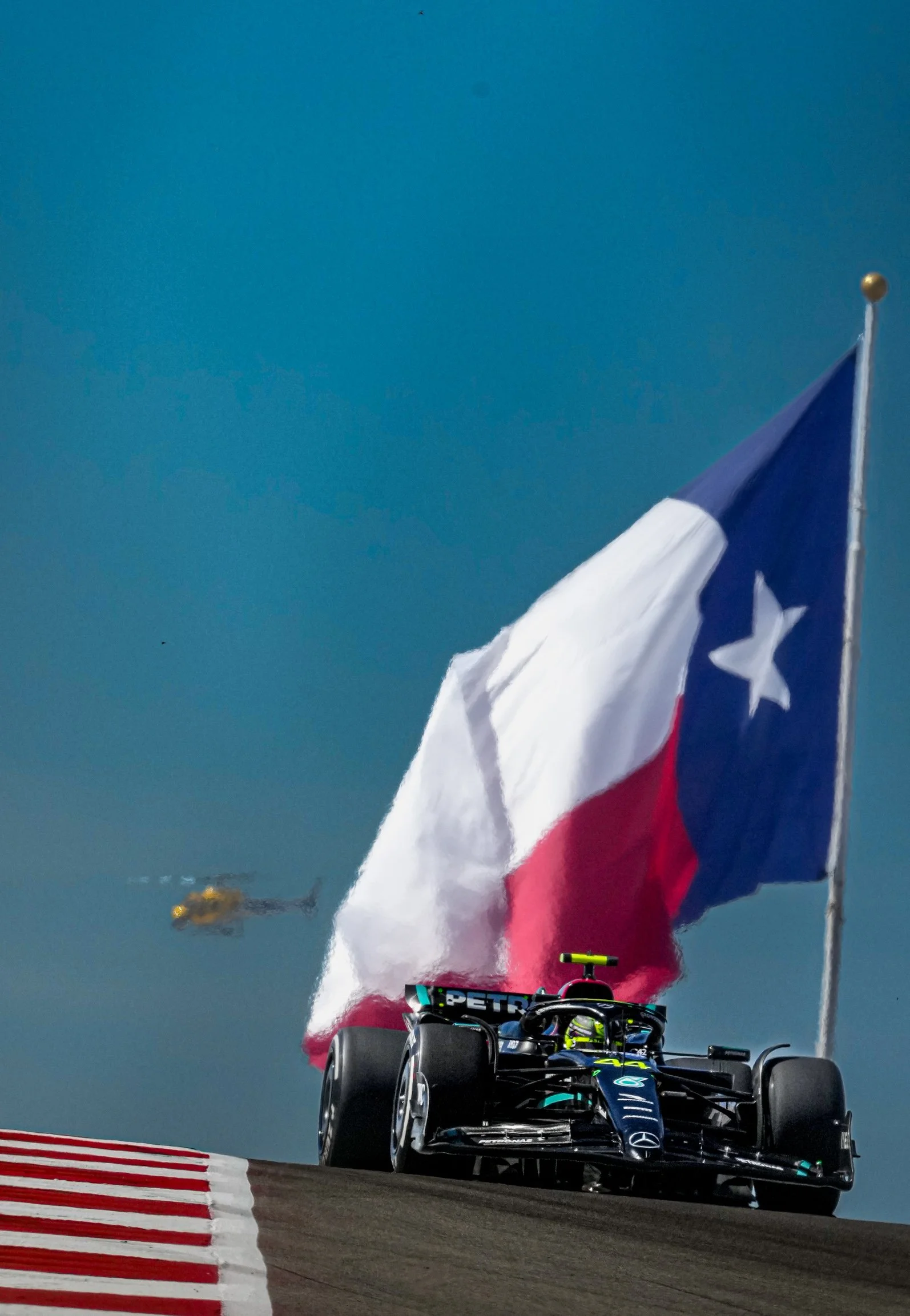 Formula 1 race car on track with Texas flag in background and helicopter in sky.