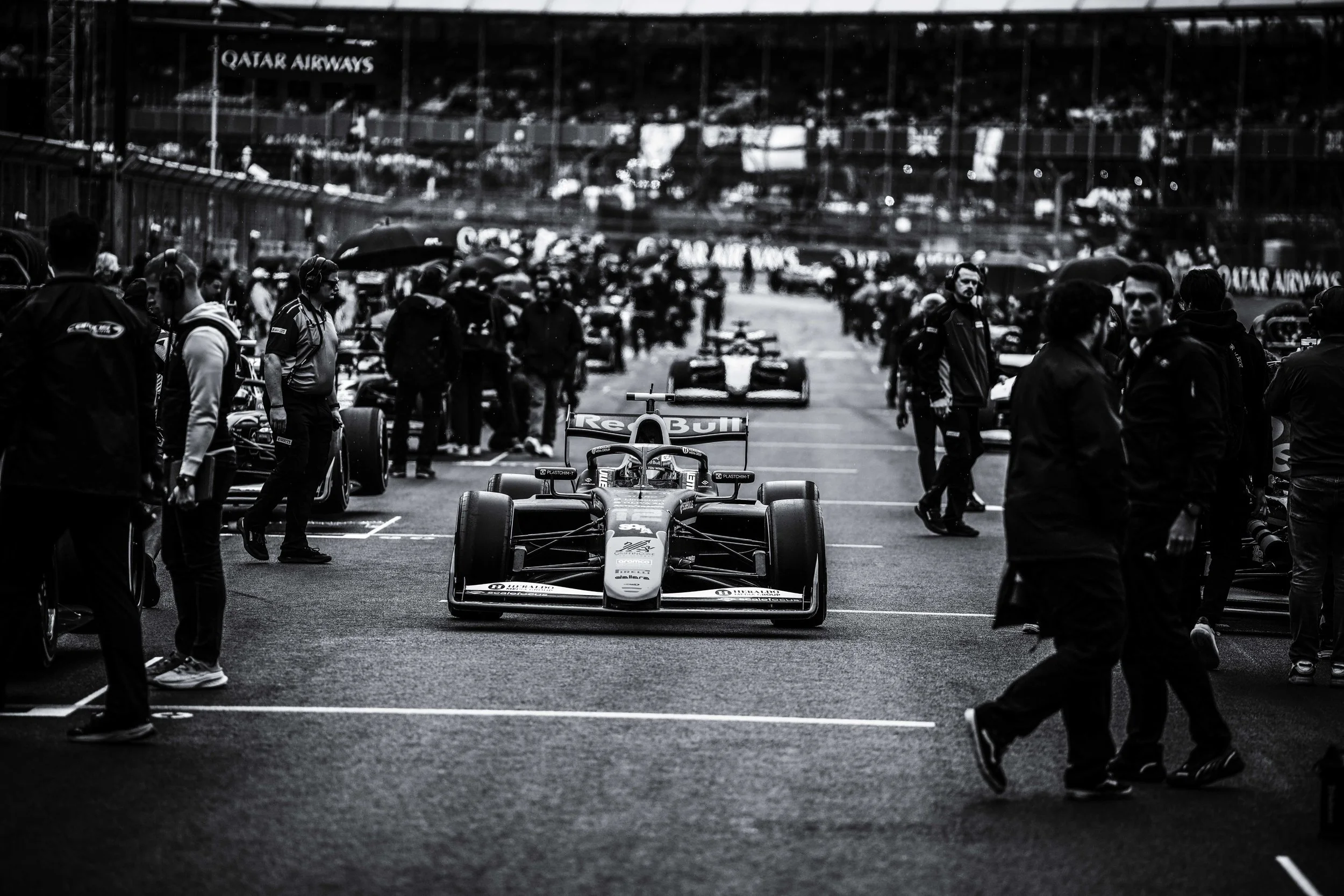Black and white photo of a Formula 1 race car in the pit lane with team members and crew around, and more racing cars in the background at a racing event.