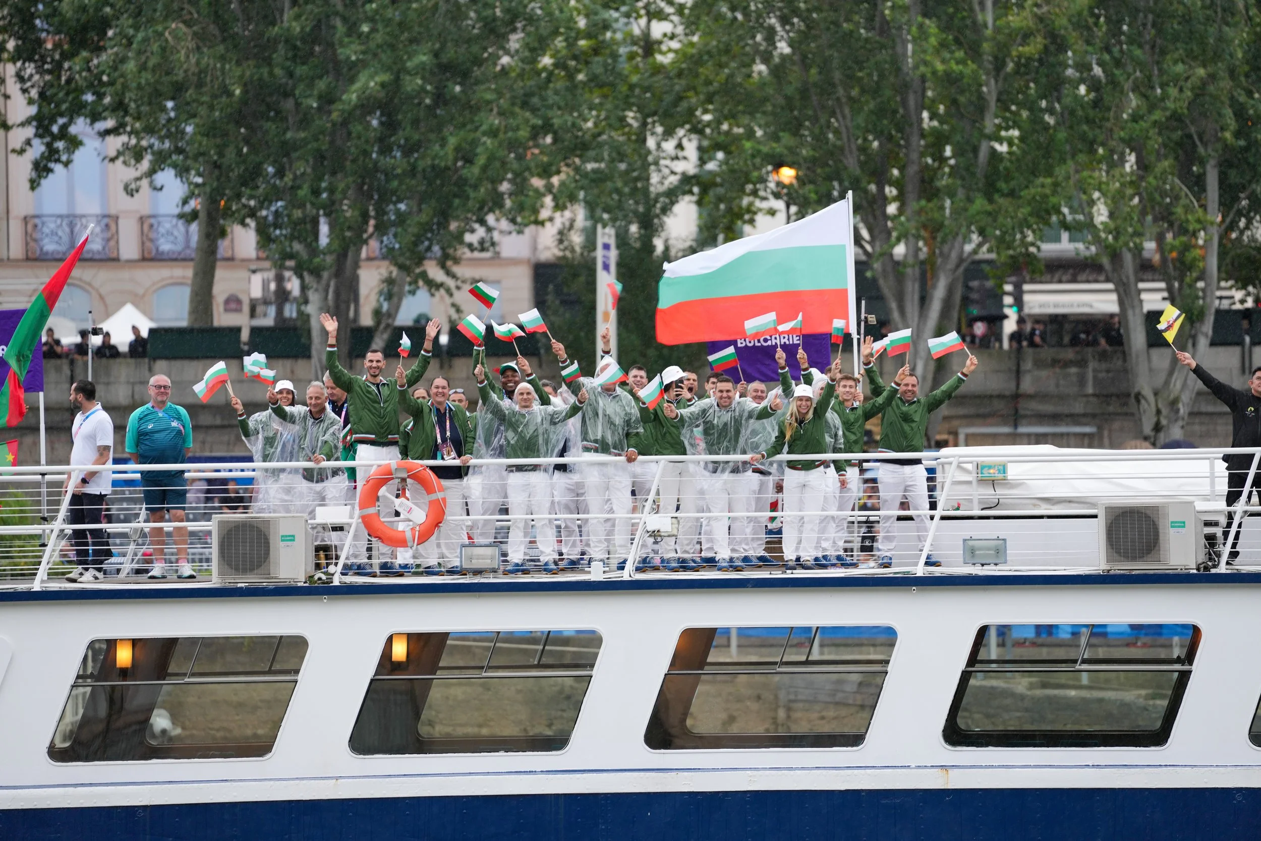 People celebrating on a boat with Bulgarian flags during a parade or event, some wearing rain ponchos, with trees and buildings in the background.