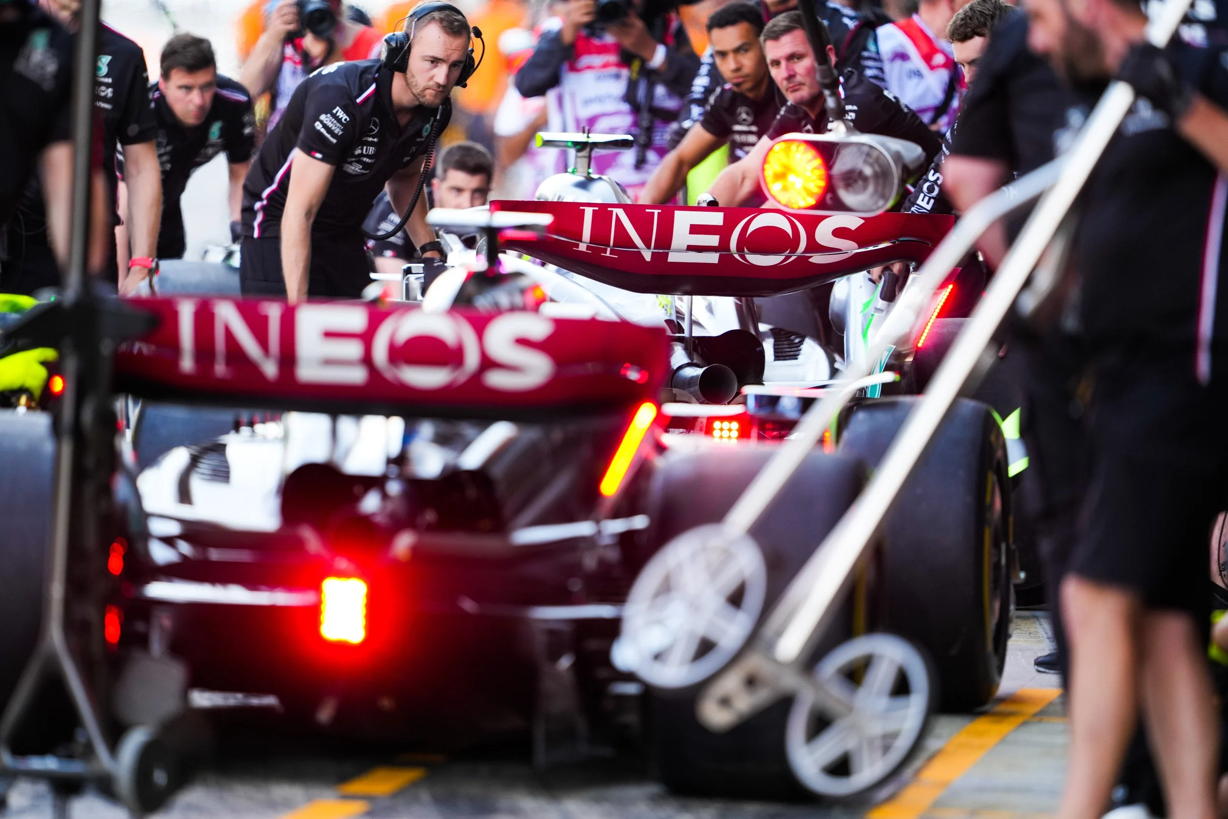 Formula 1 cars in the pit lane with team members working on them during a race, with one car displaying the 'INEOS' sponsor logo.