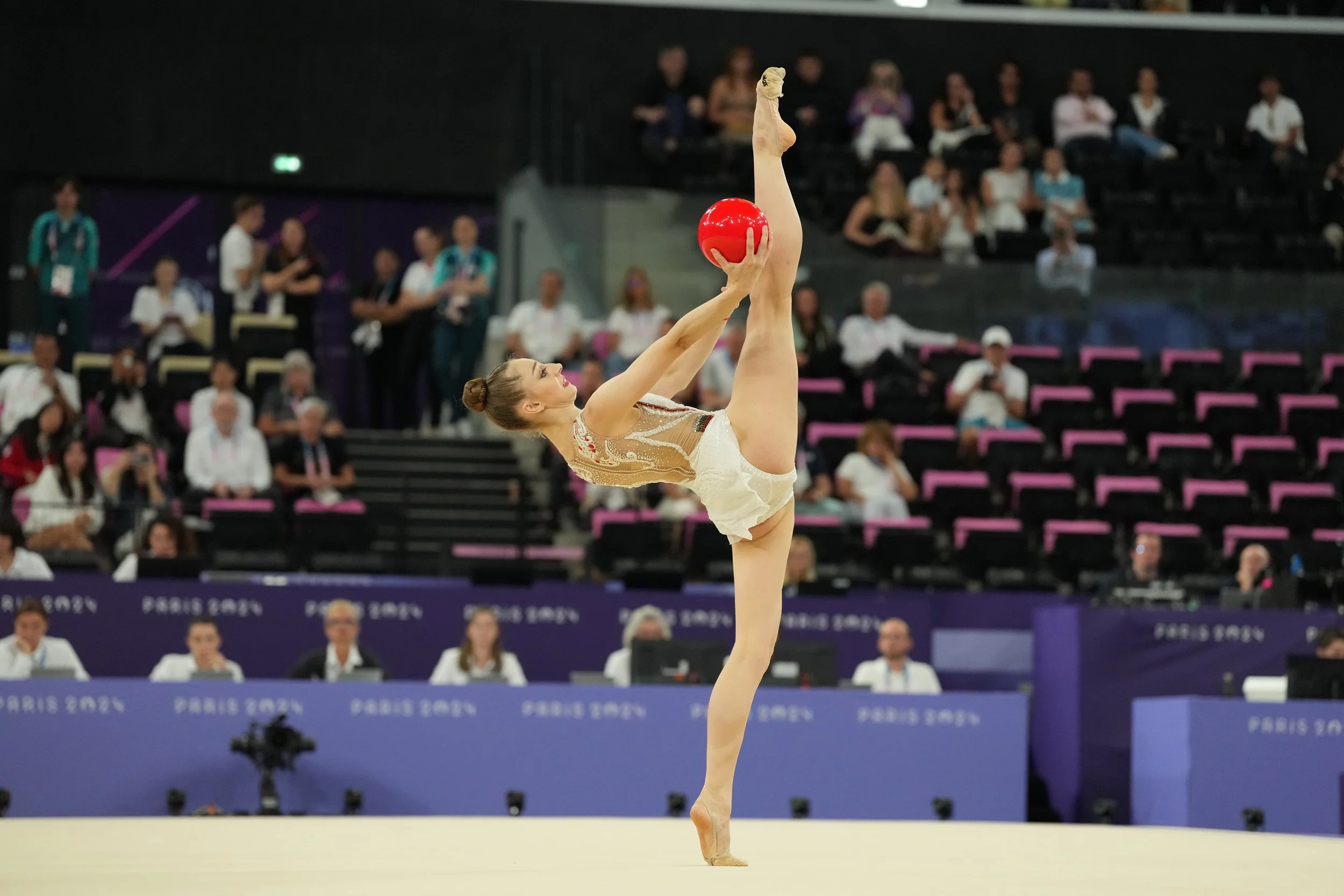 A female gymnast performing a balance pose with one leg raised vertically while holding a red ball during a gymnastics competition at the Paris 2024 Olympic Games.