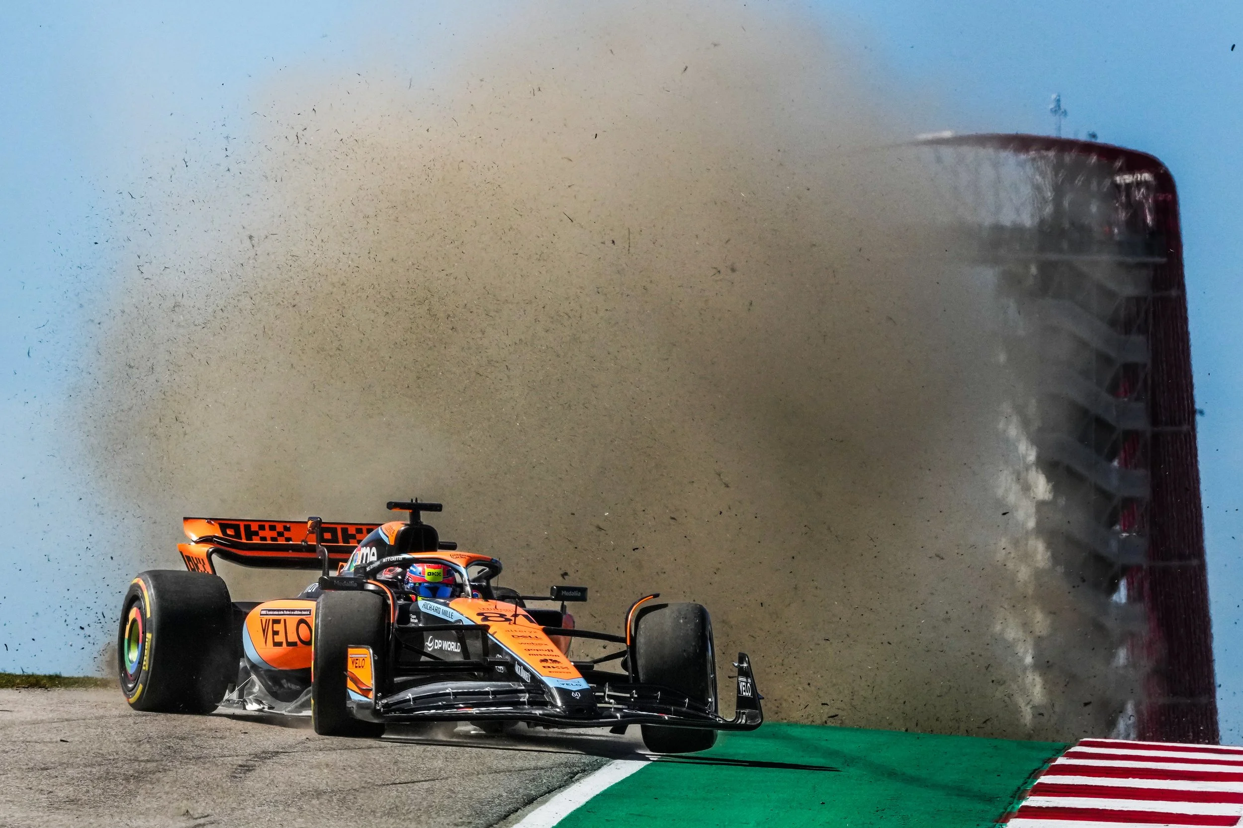 An orange and black IndyCar racing car speeding on a track, kicking up dirt and dust with a group of spectators visible in the background.