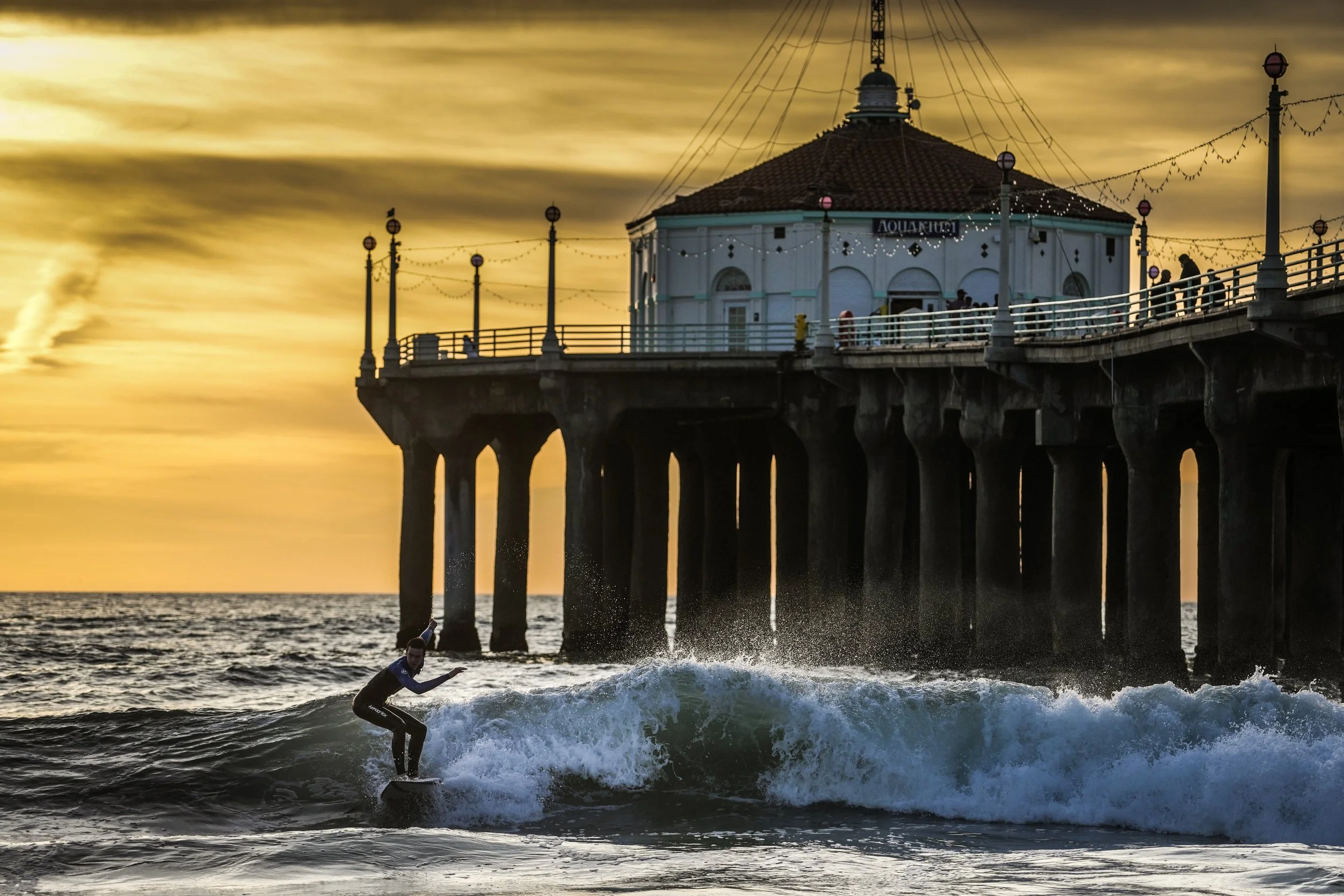 A surfer riding a wave near a pier at sunset, with a building on the pier and oil refinery smoke in the background.