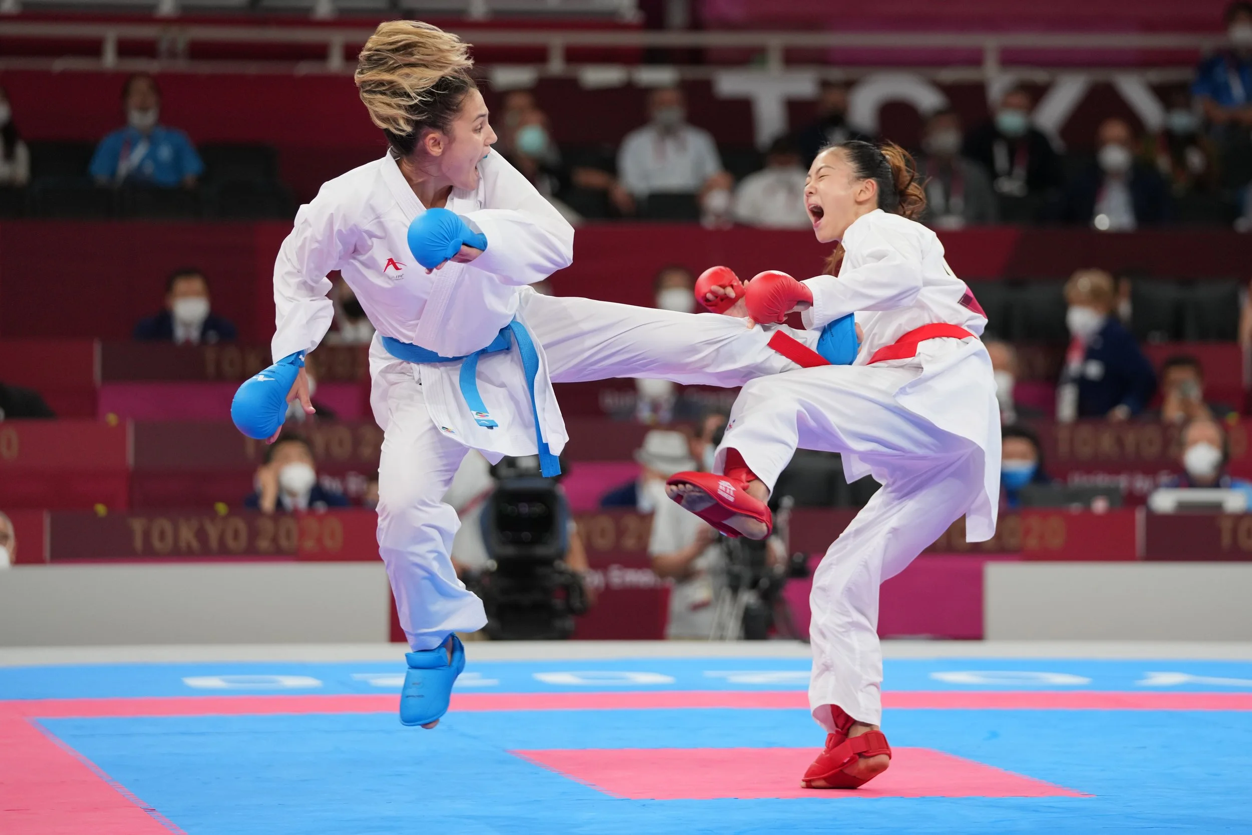 Two female martial artists competing in a karate match at Tokyo 2020, both wearing traditional white uniforms and protective gloves, executing a high kick toward each other on the mat, with spectators in the background.