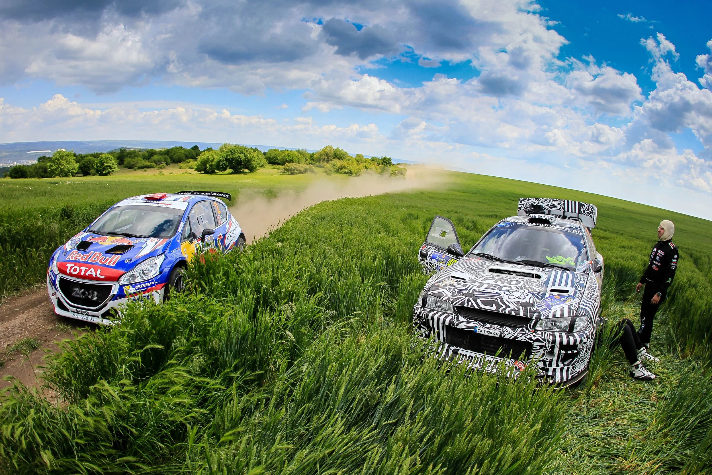 Two rally cars are parked in a grassy field with a person standing nearby. One vehicle is momentarily off-road in tall grass, and the other is partially hidden by grass. The background features a blue sky with scattered clouds and a distant green lan