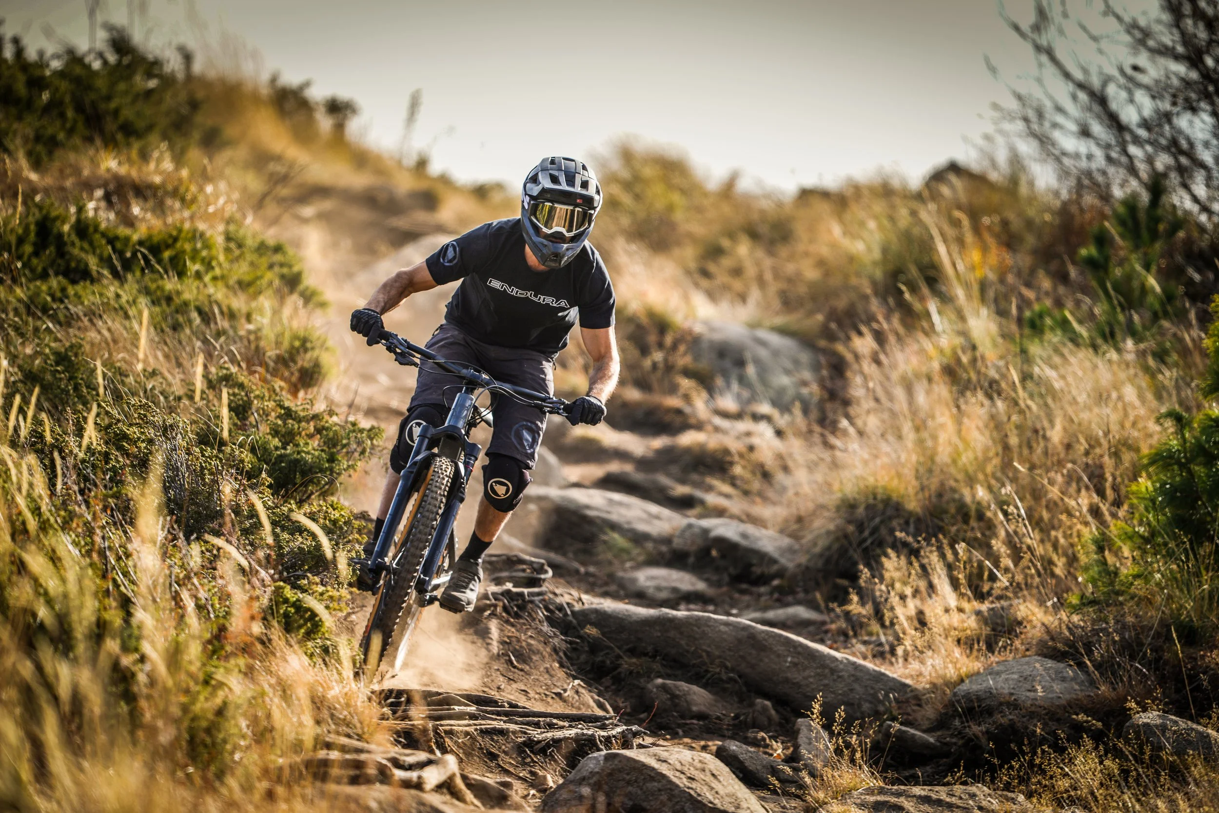 A mountain biker wearing a helmet and gloves rides on a rocky, dirt trail surrounded by dry grass and bushes.