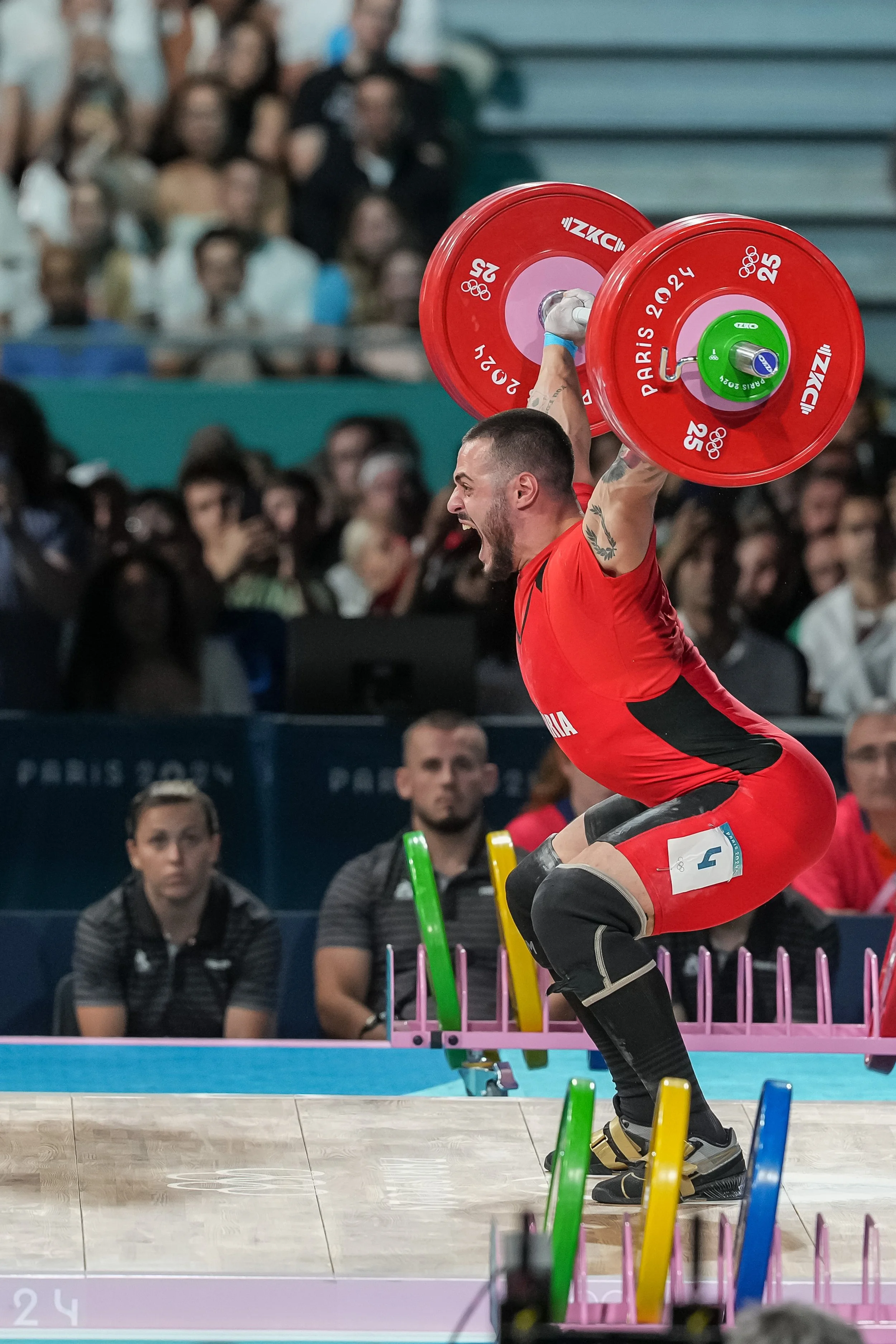 A male weightlifter in red uniform lifts a barbell above his head during a competition at Paris 2024. Spectators watch from the stands.