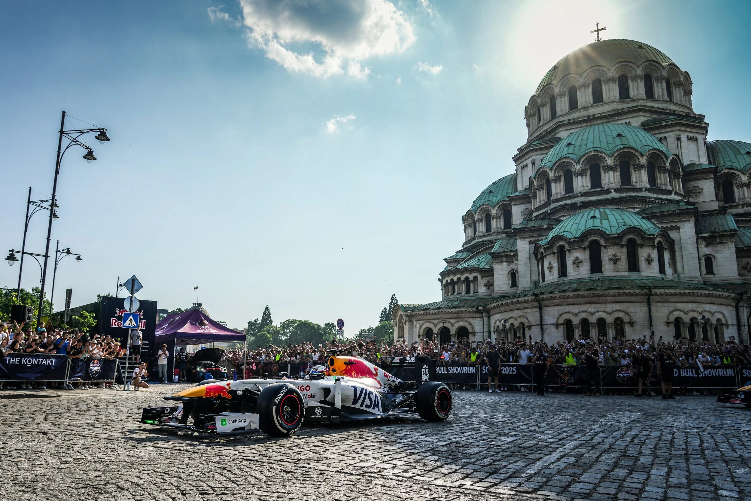A Formula 1 race car driving on cobblestone street in front of a large crowd and a historic church with domes and a cross on top, under a partly cloudy sky.