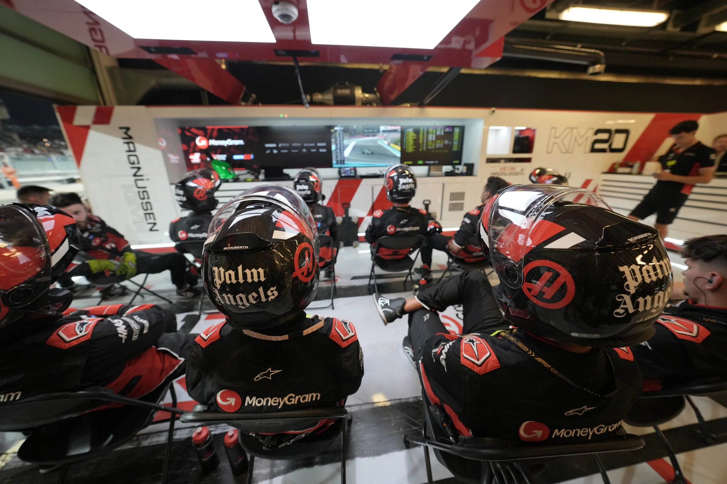 Formula 1 race team members in racing suits and helmets, seated in front of a large screen monitoring race data, in a racing garage setting.