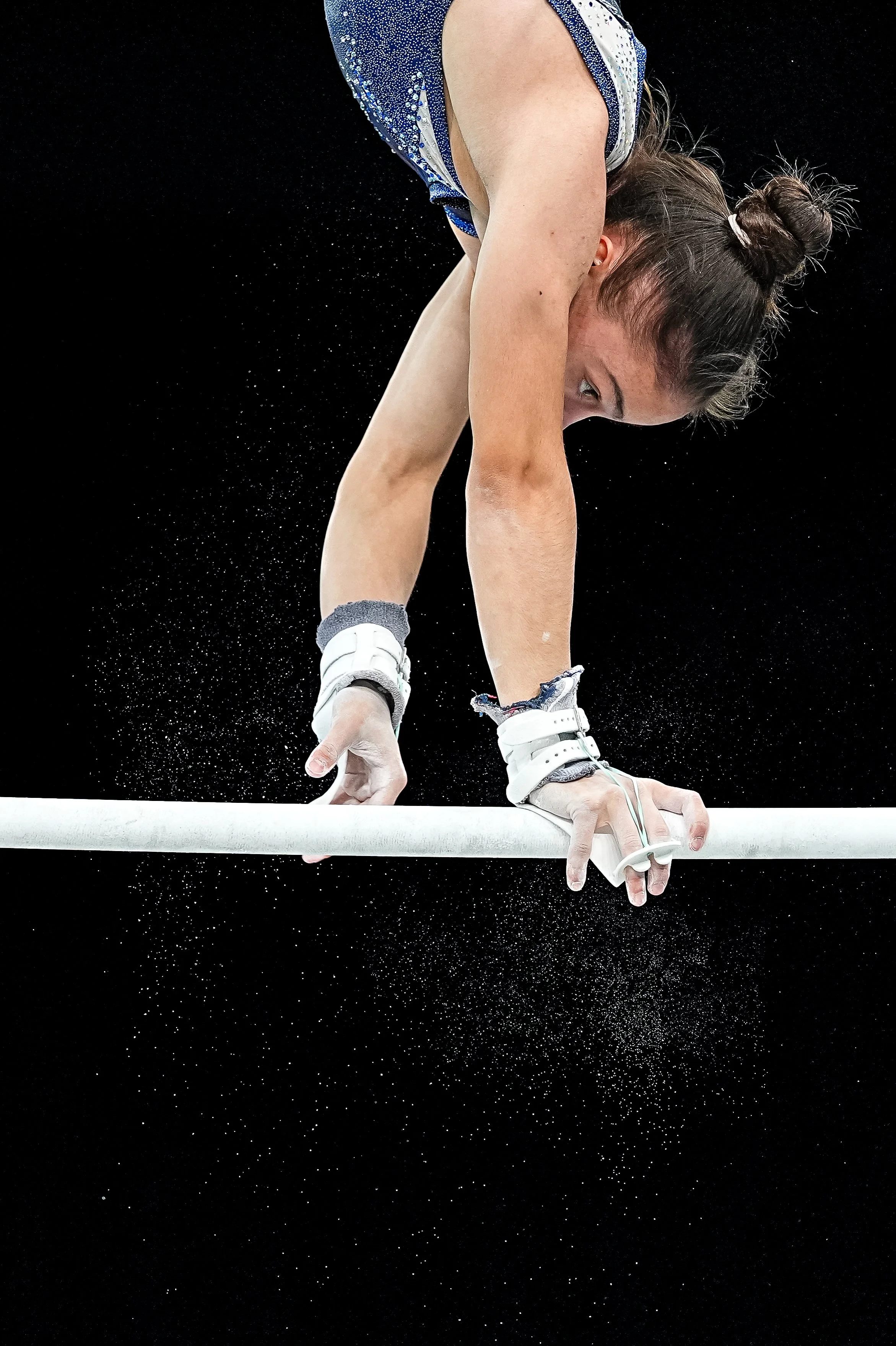 A female gymnast in a blue and white leotard performing on the uneven bars, with chalk dust in the air against a dark background.