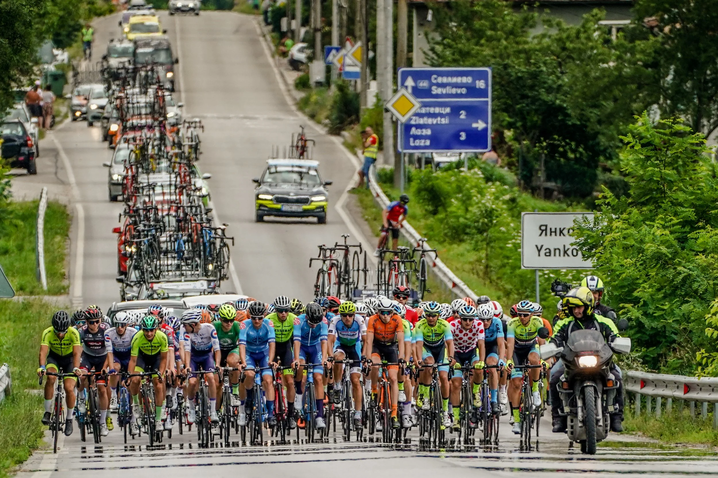 A group of cyclists participating in a race on a paved road with green trees on the side, accompanied by race officials and support motorcycles, and cars with bicycles on a rack in the background.