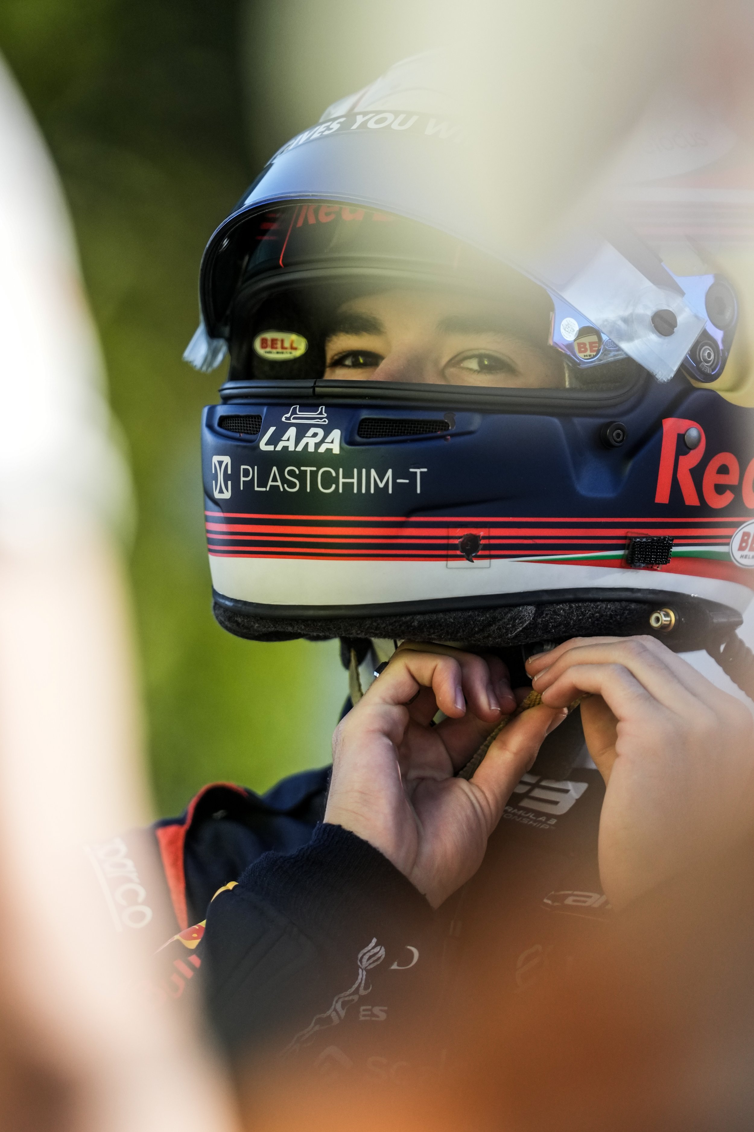 A race car driver wearing a helmet with sponsors' labels, including Red Bull and Bell, adjusting the strap of his helmet during a race or practice session.