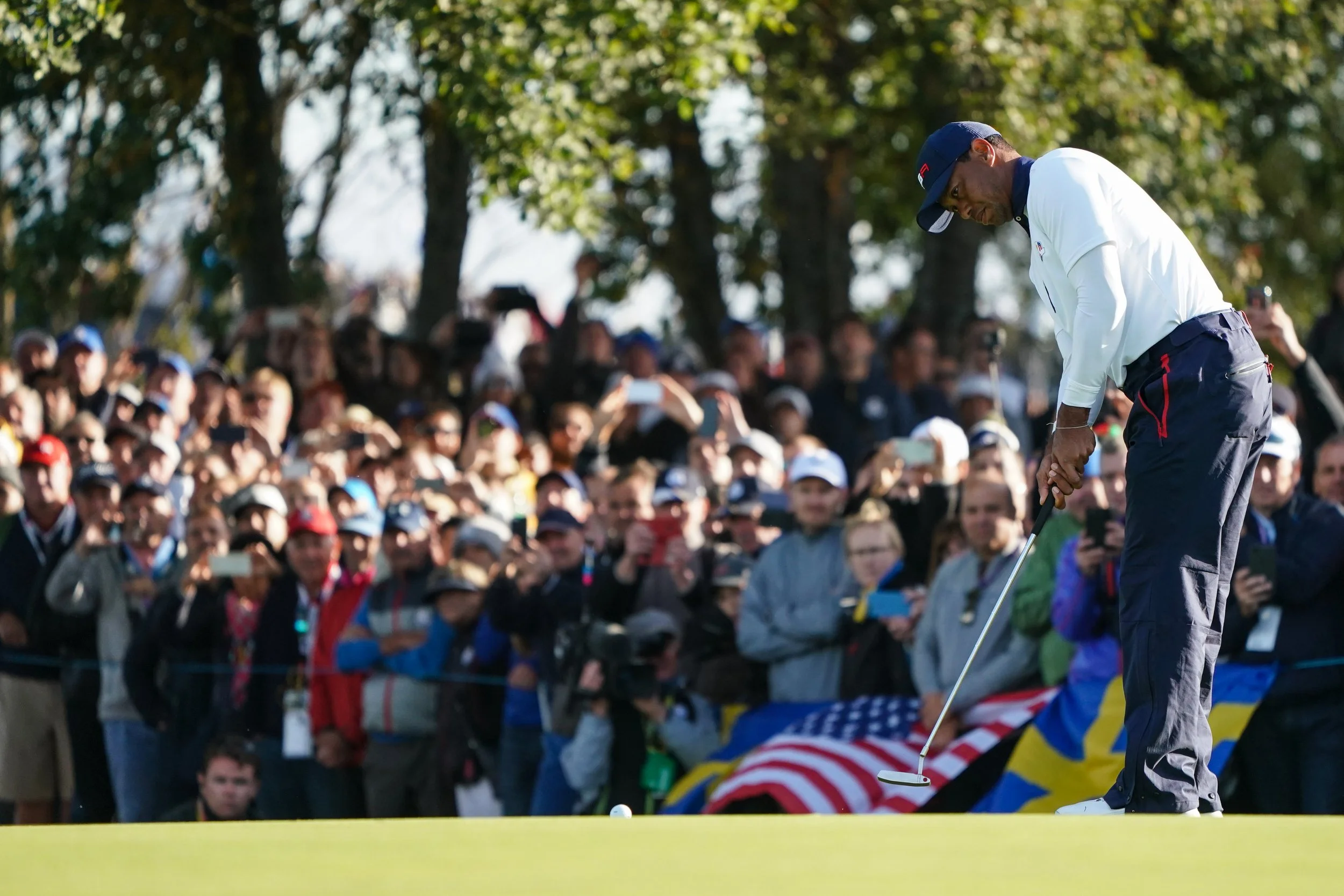 A golfer lining up a putt on the green while a crowd of spectators watches behind a rope, holding phones and cameras. Trees are visible in the background.