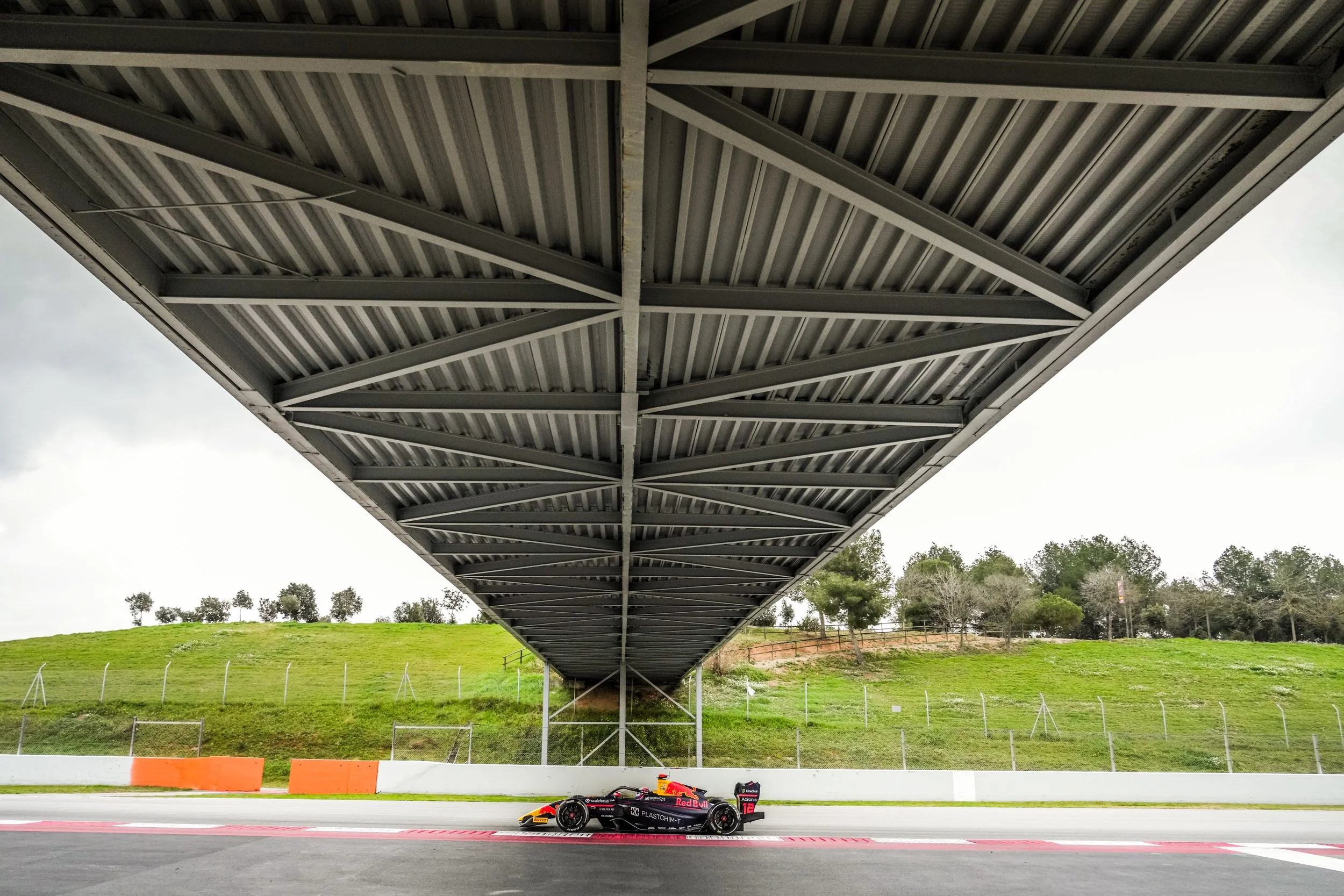 A Formula 1 race car with Red Bull branding parked on a race track beneath a large metal overpass, with green grassy hills and trees in the background.