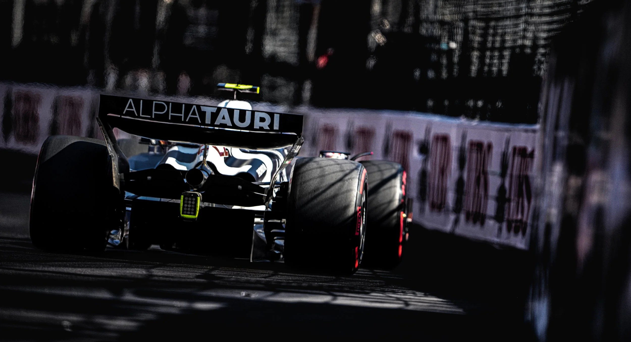 A Formula 1 race car on a dark track with shadowed areas, featuring a rear wing with the Alfa Tauri branding, large slick tires, and a blurred background with barriers.