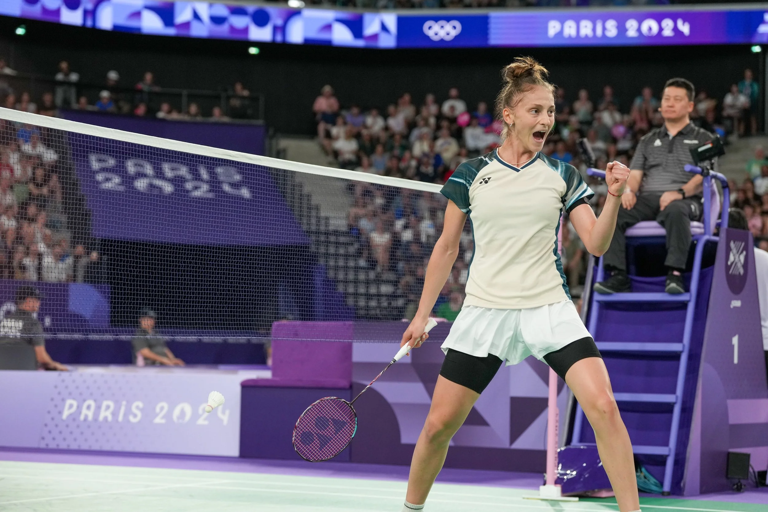 A female badminton player celebrates on the court at the Paris 2024 Olympics, holding a racket, with a crowd and officials in the background.