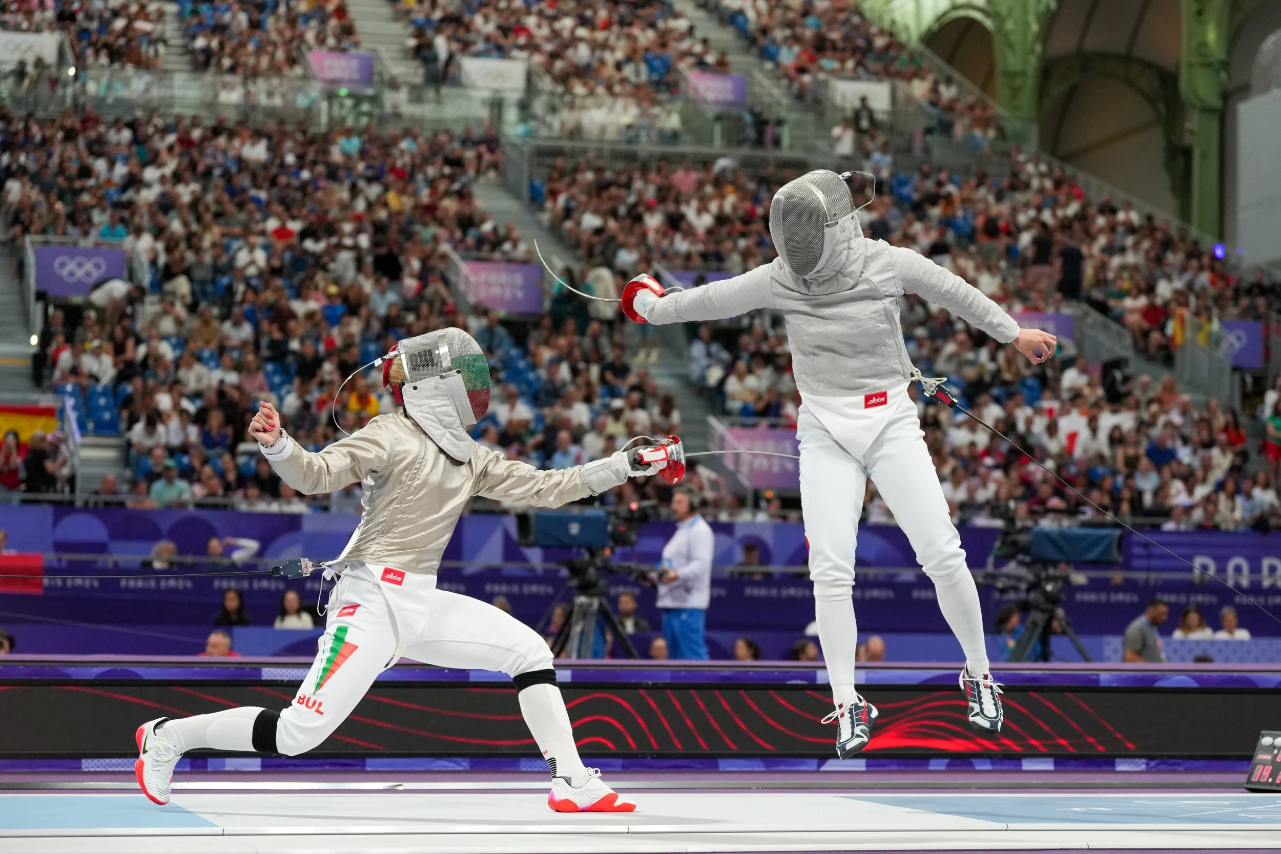 Two Olympic fencers, one in mid-air lunging at the other, both wearing protective gear and helmets, during a match at the Olympics with a large crowd in the background.