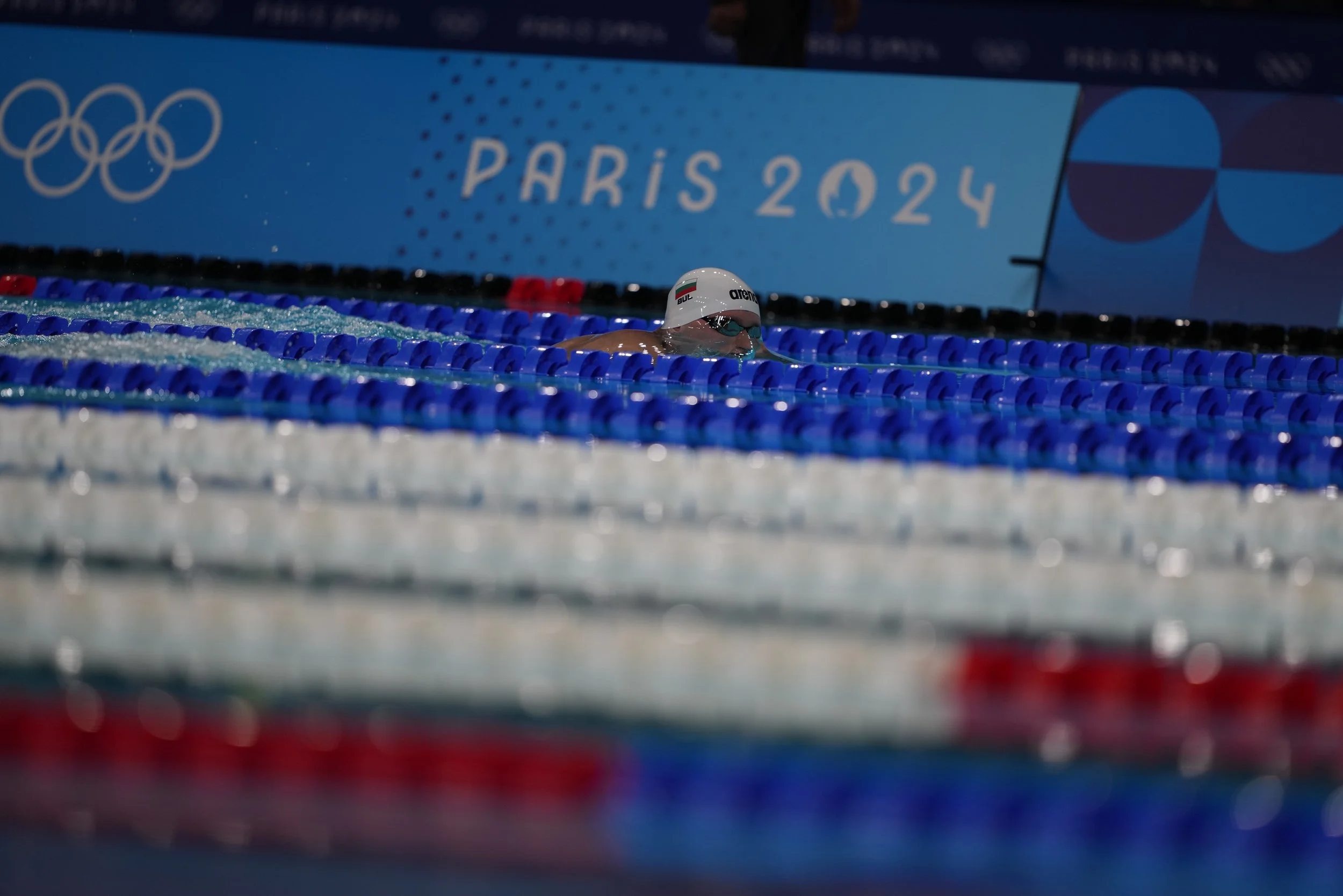 A swimmer in a pool during the Paris 2024 Olympics, wearing goggles and a swim cap, swimming towards the wall.