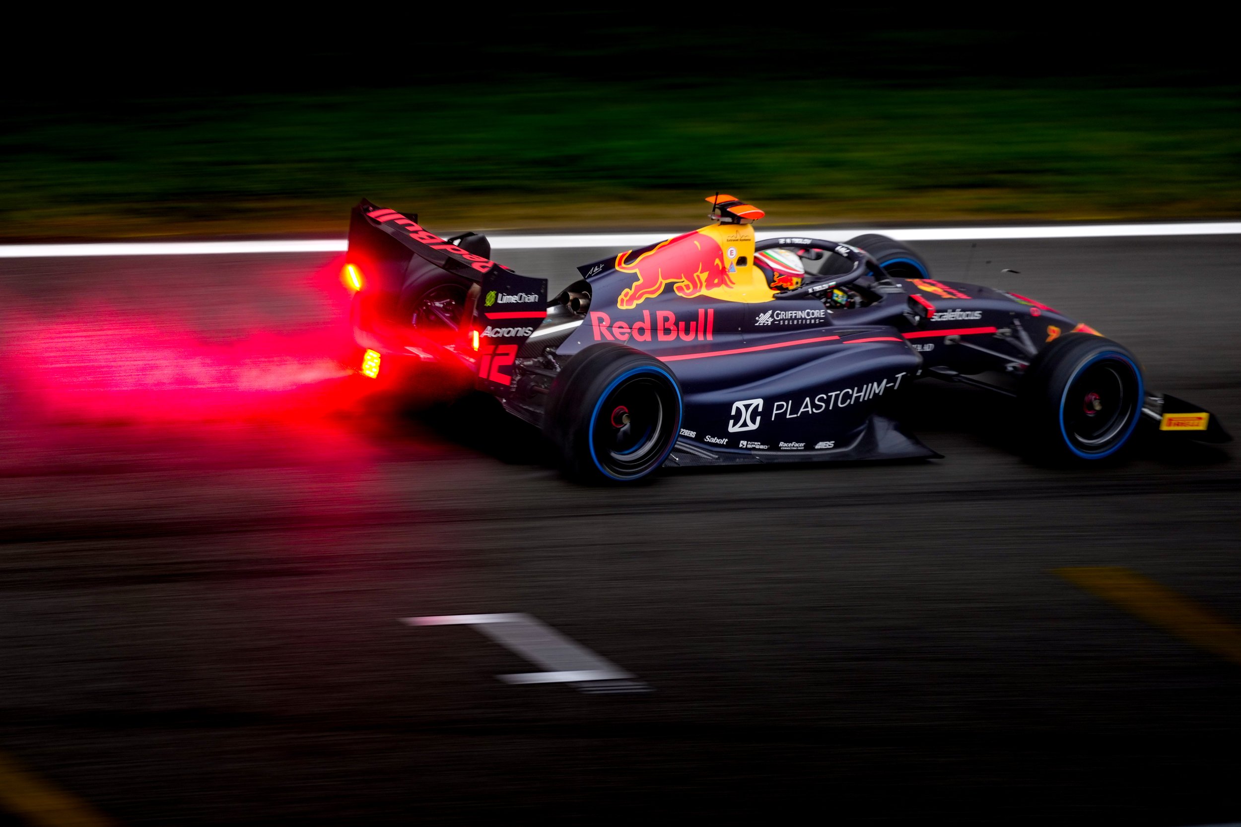 A dark blue Formula 2 race car with Red Bull branding racing on a track, emitting red flame from the exhaust.