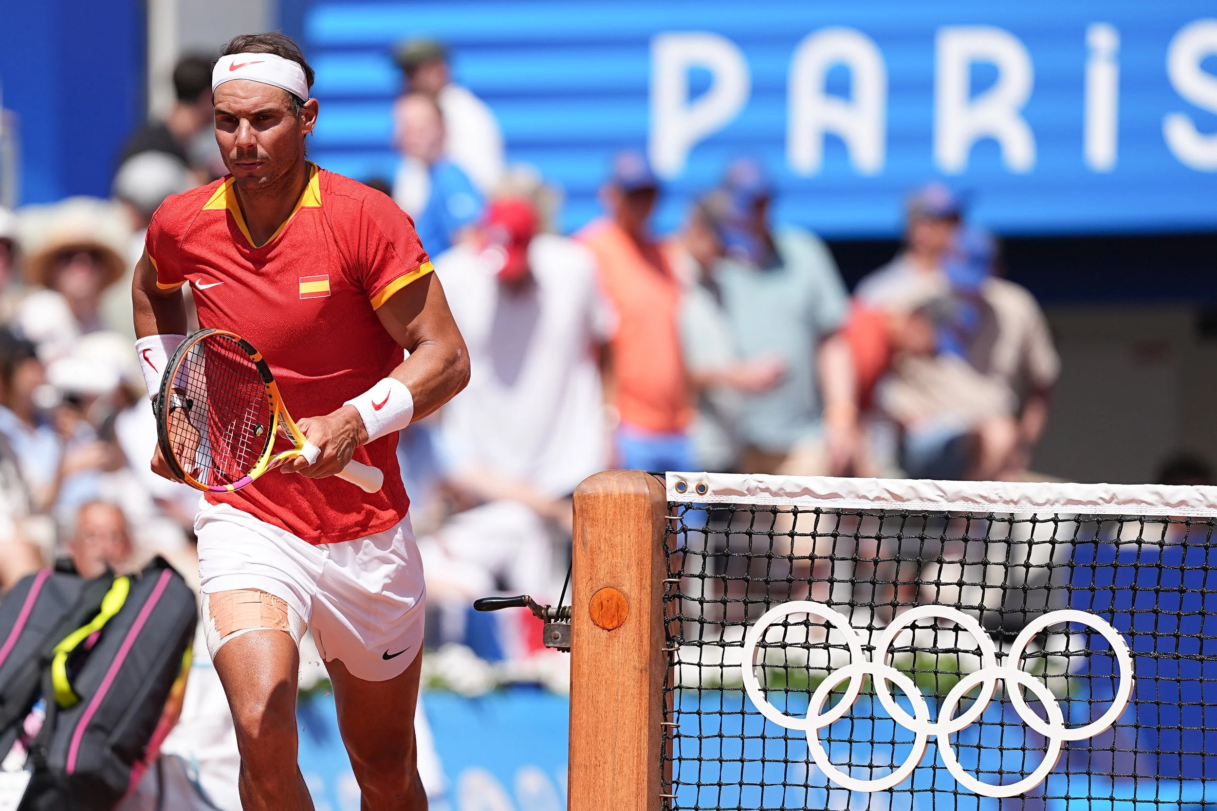 A male tennis player in a red shirt and white shorts, holding a tennis racket, near a tennis net with the Olympic rings, during a match at the Tokyo Olympics.