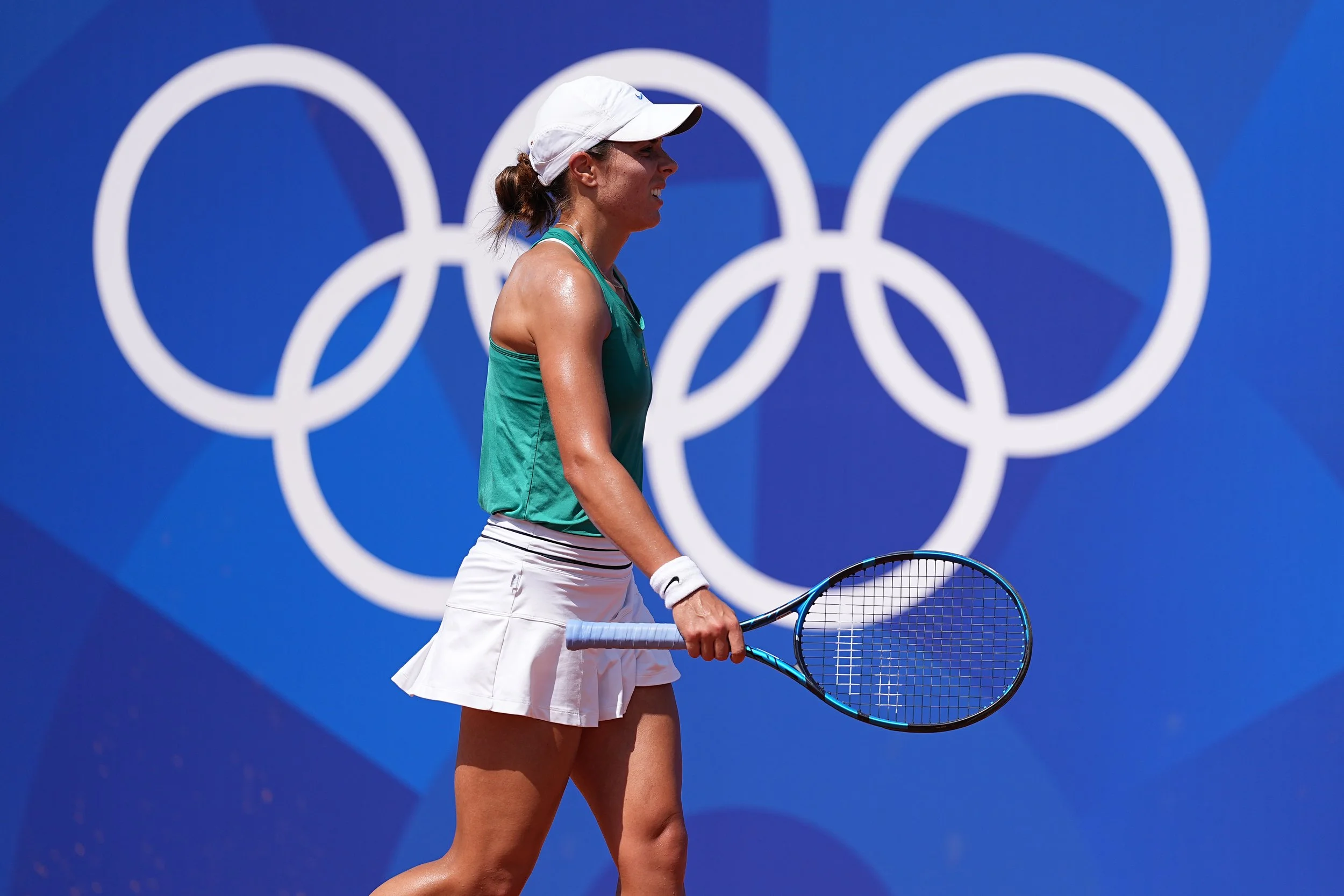 A female tennis player in green and white outfit holding a racket with a blue background featuring the Olympic rings.