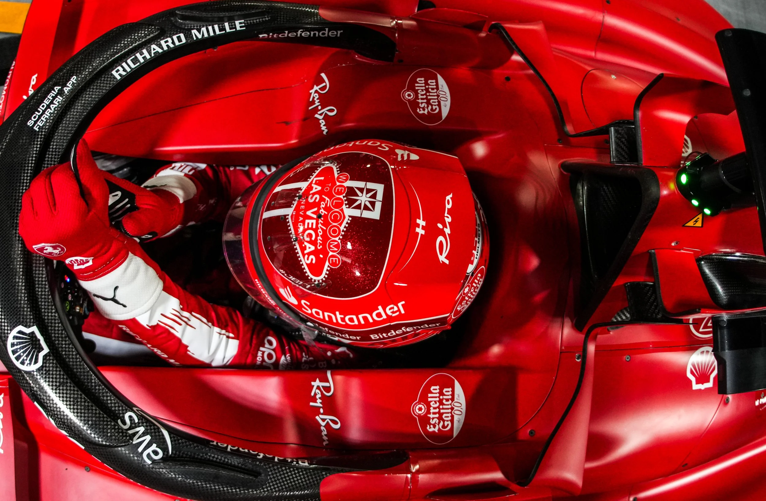 Top-down view of a Ferrari Formula 1 race car driver in a red racing helmet and suit, seated in the cockpit surrounded by red bodywork with sponsorship logos, including Shell, Santander, and Estrella Galicia.