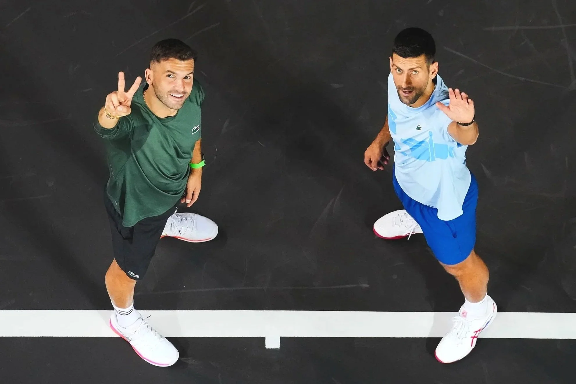 Grigor Dimitrov and Novak Jokovic stand on a tennis court, looking up at the camera, waving and smiling.
