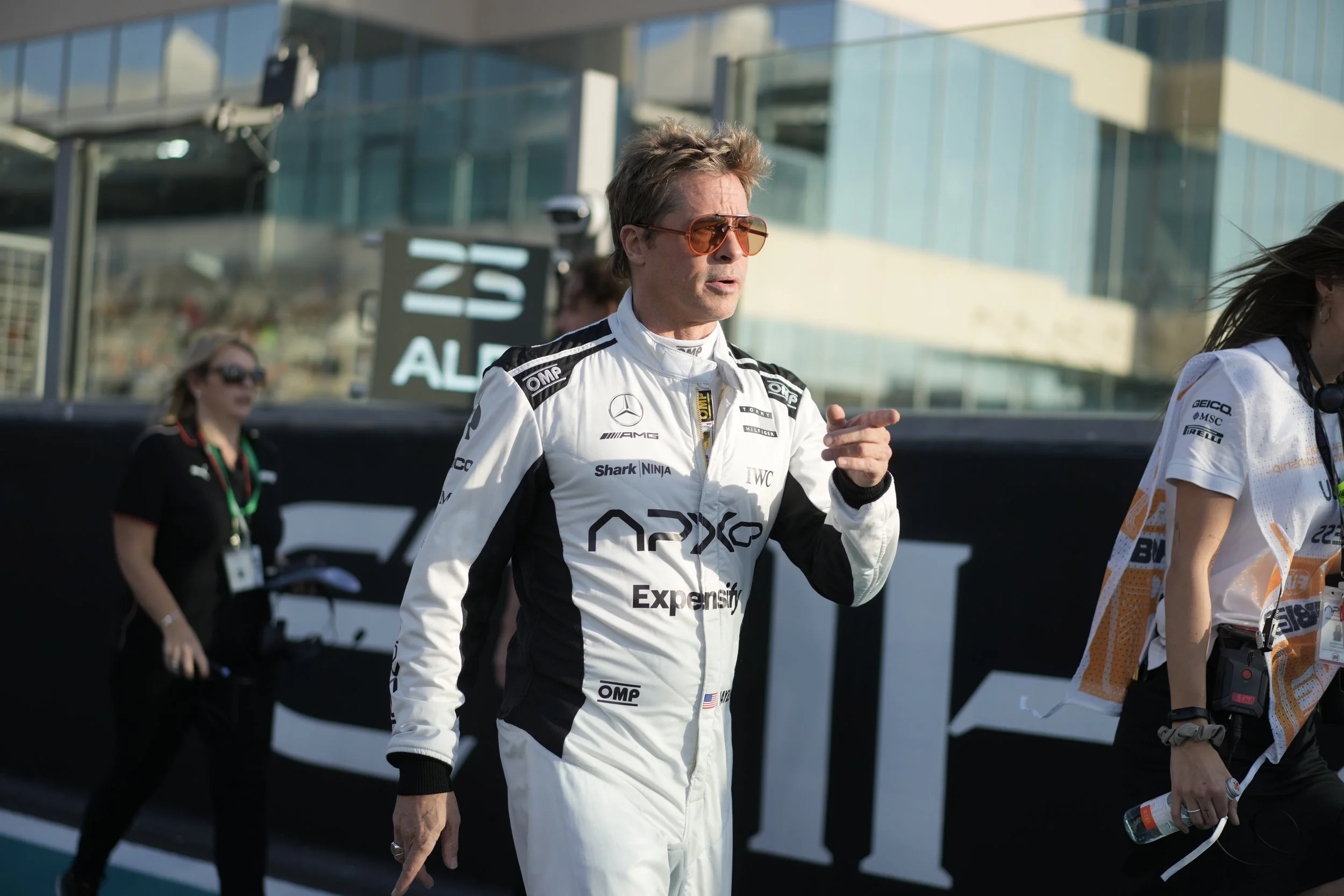 Man in a racing suit with sunglasses standing on a racing circuit, with people around him and modern buildings in the background.