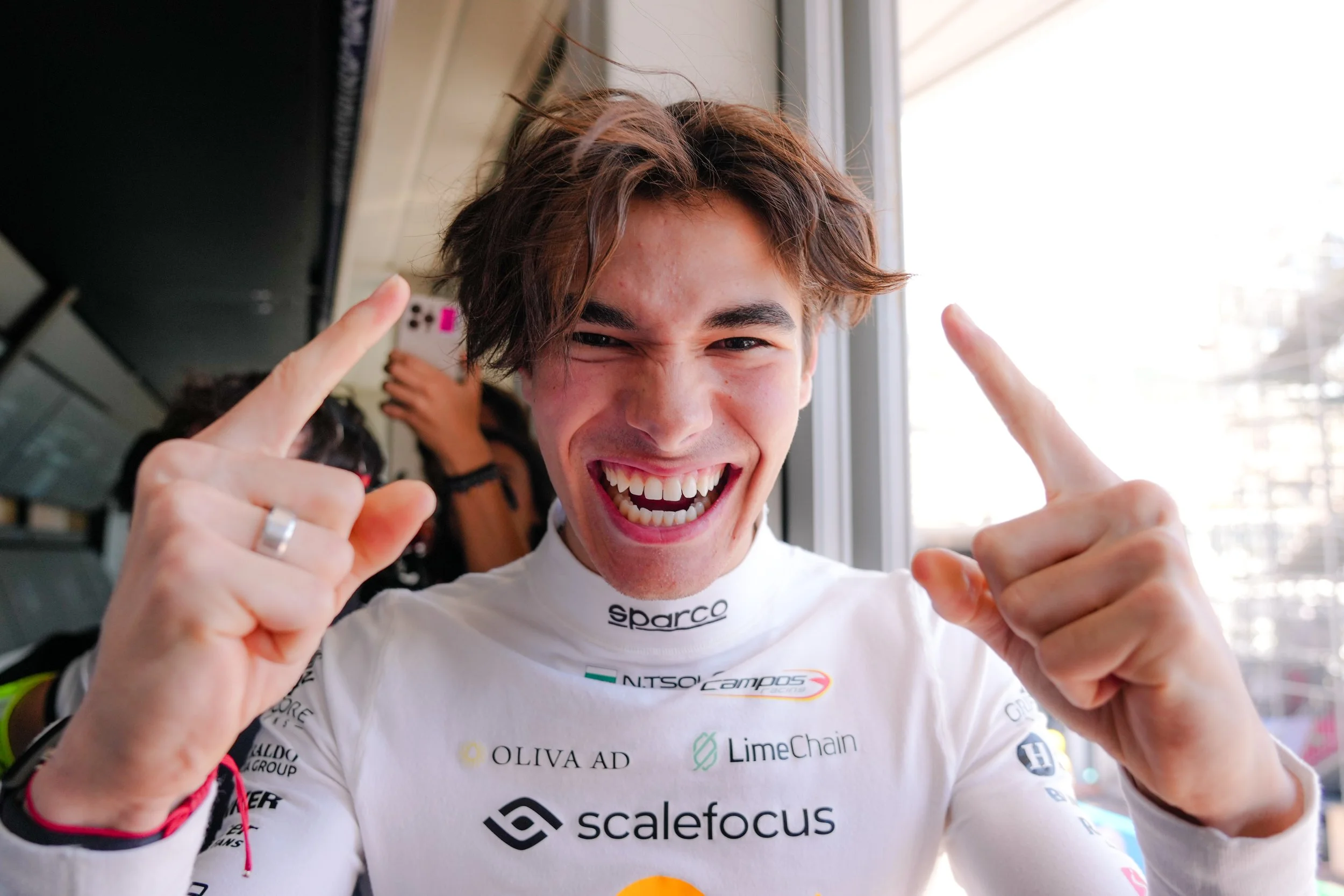 A young man with tousled brown hair wearing a white racing suit, smiling widely, pointing upwards with both index fingers.
