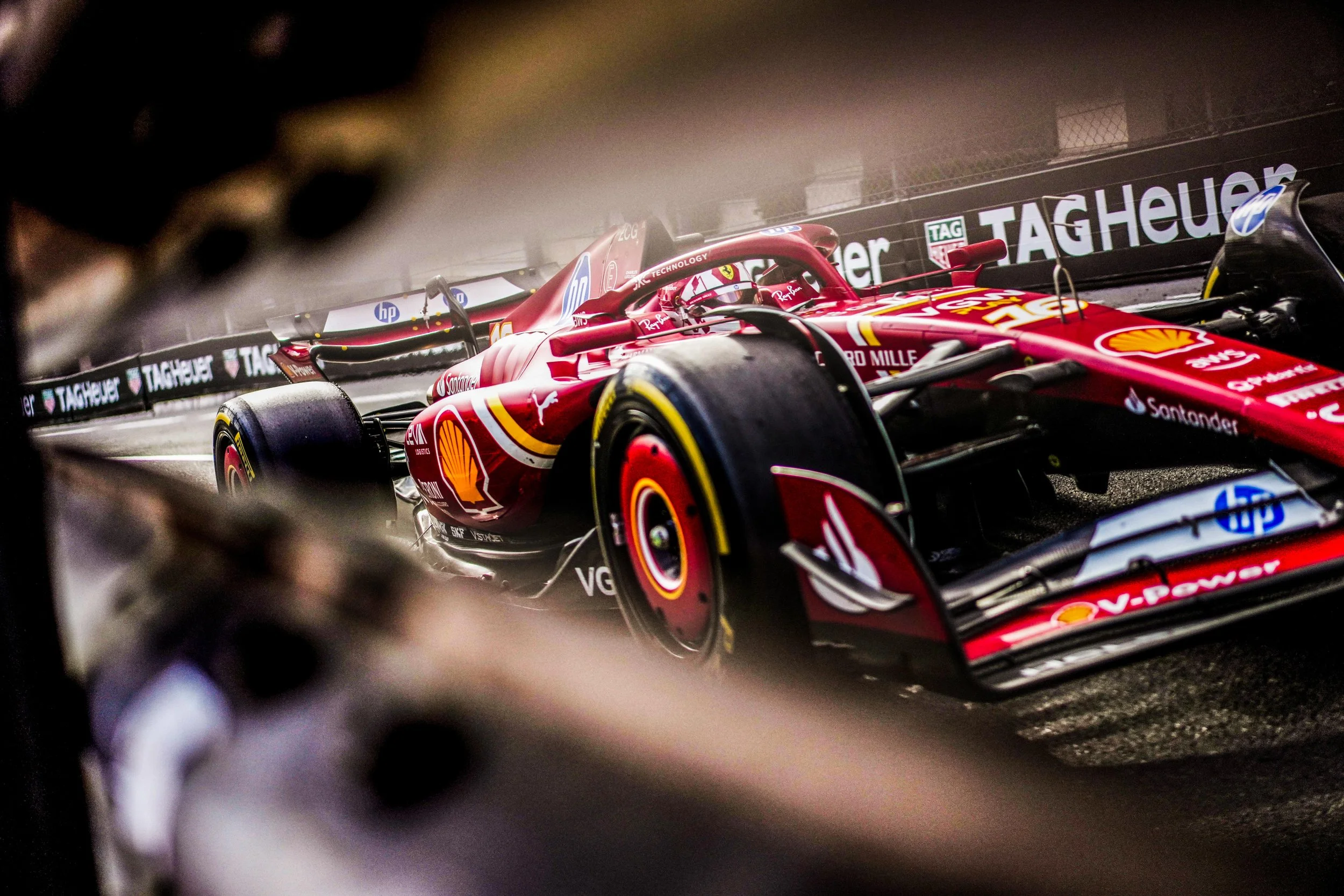 A red Formula 1 race car on a race track, viewed through a blurred opening, with sponsorship logos including Shell, Shell V-Power, Santander, SAP, HP, and TAG Heuer visible on the car and track barriers.