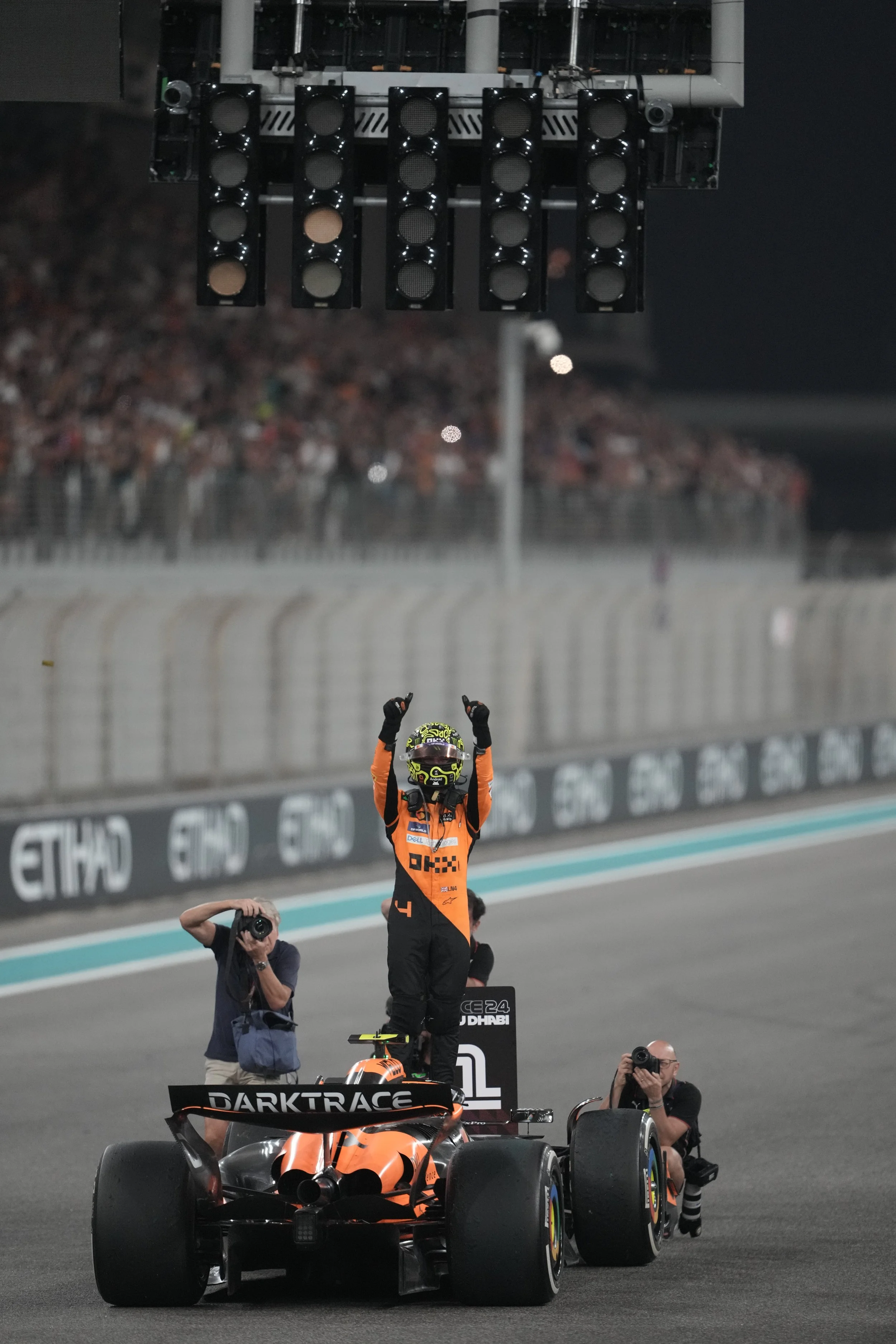 A race car driver standing on the wing of his race car, celebrating victory with raised arms, while photographers capture the moment on the track at night.