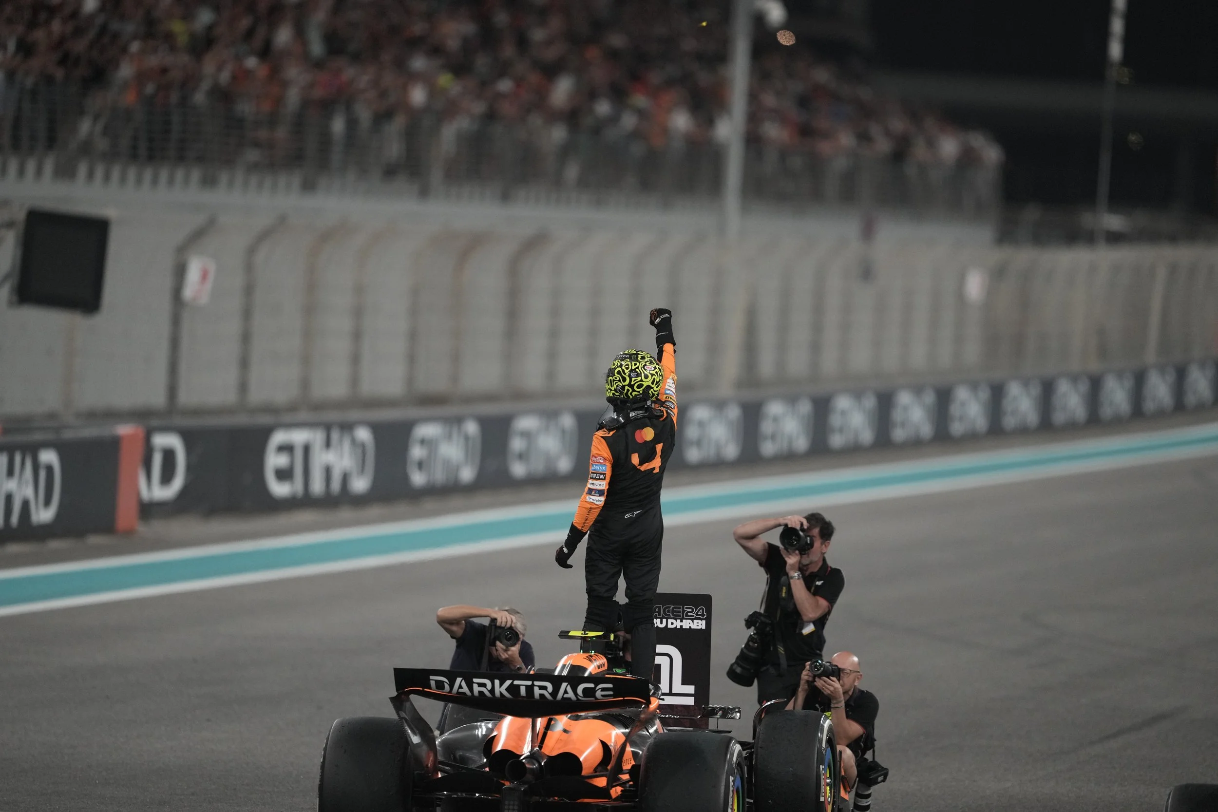Race car driver in racing suit and helmet standing on top of a race car, raising an arm in victory, with photographers capturing the moment on a race track at night.