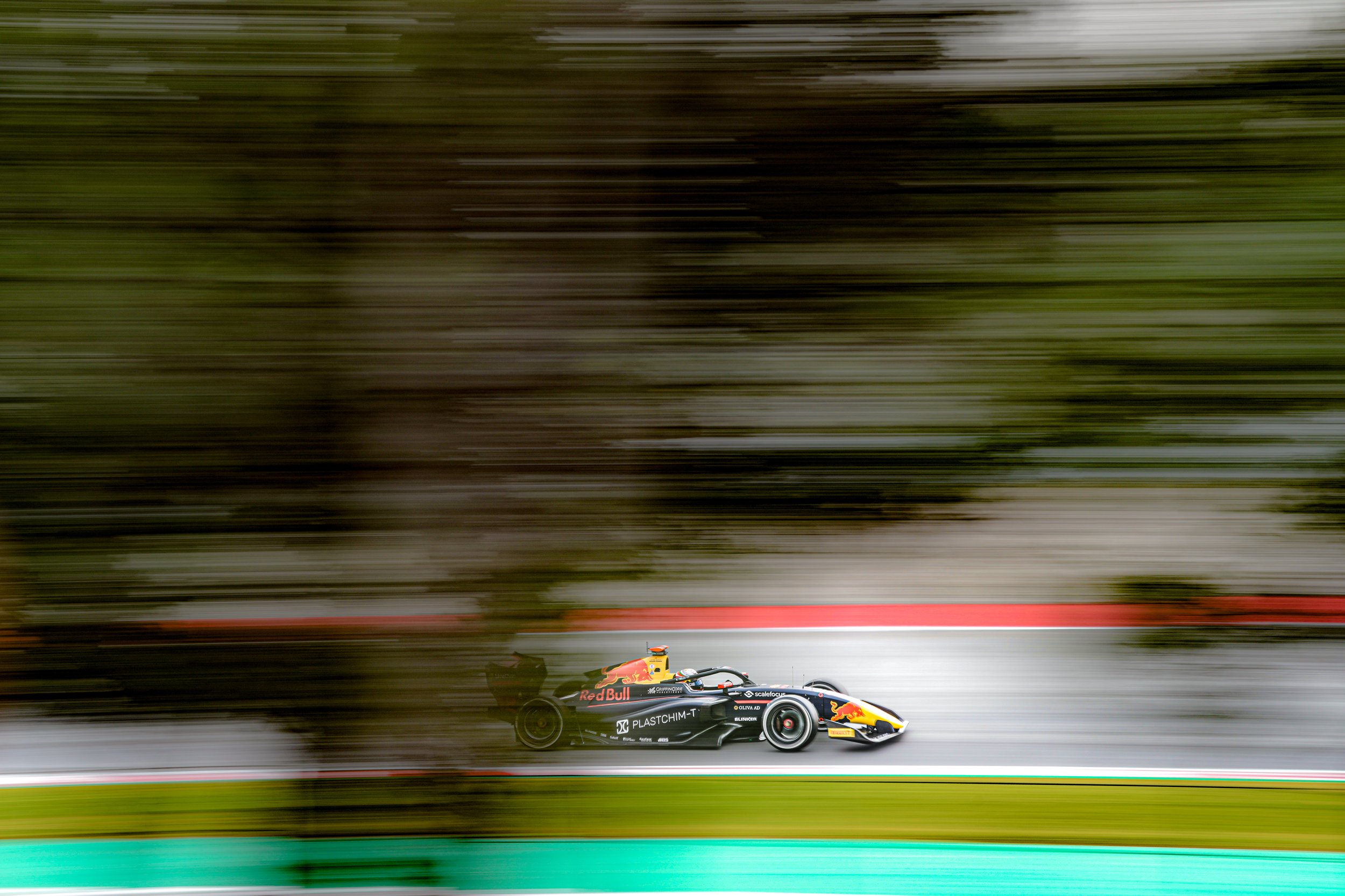 A racing car moving at high speed on a track with motion blur, trees and a section of the track in the background.